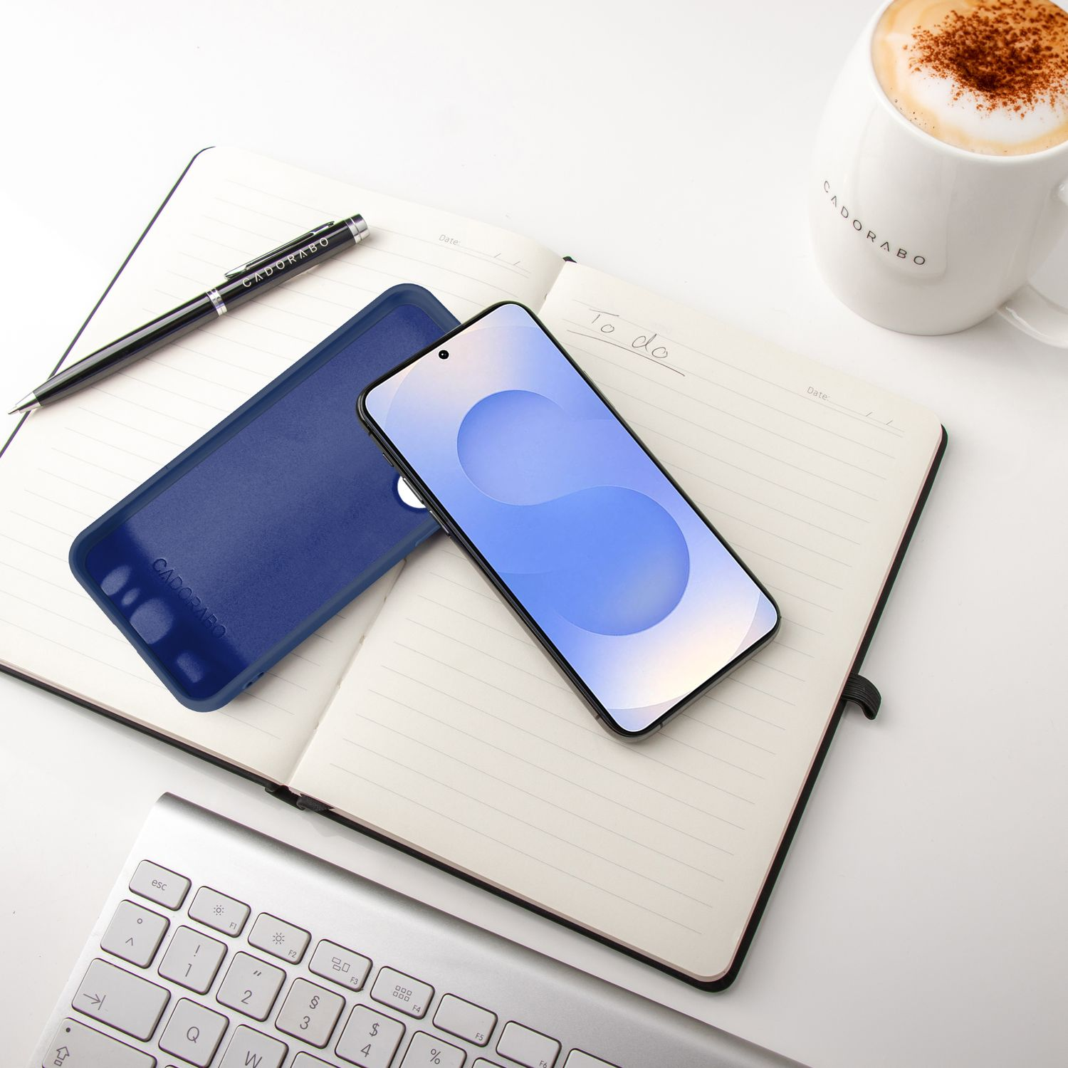 Ein Telefon, eine Hülle, ein Stift und eine Tasse Kaffee liegen auf einem Notizbuch. Im Hintergrund eine Tastatur.