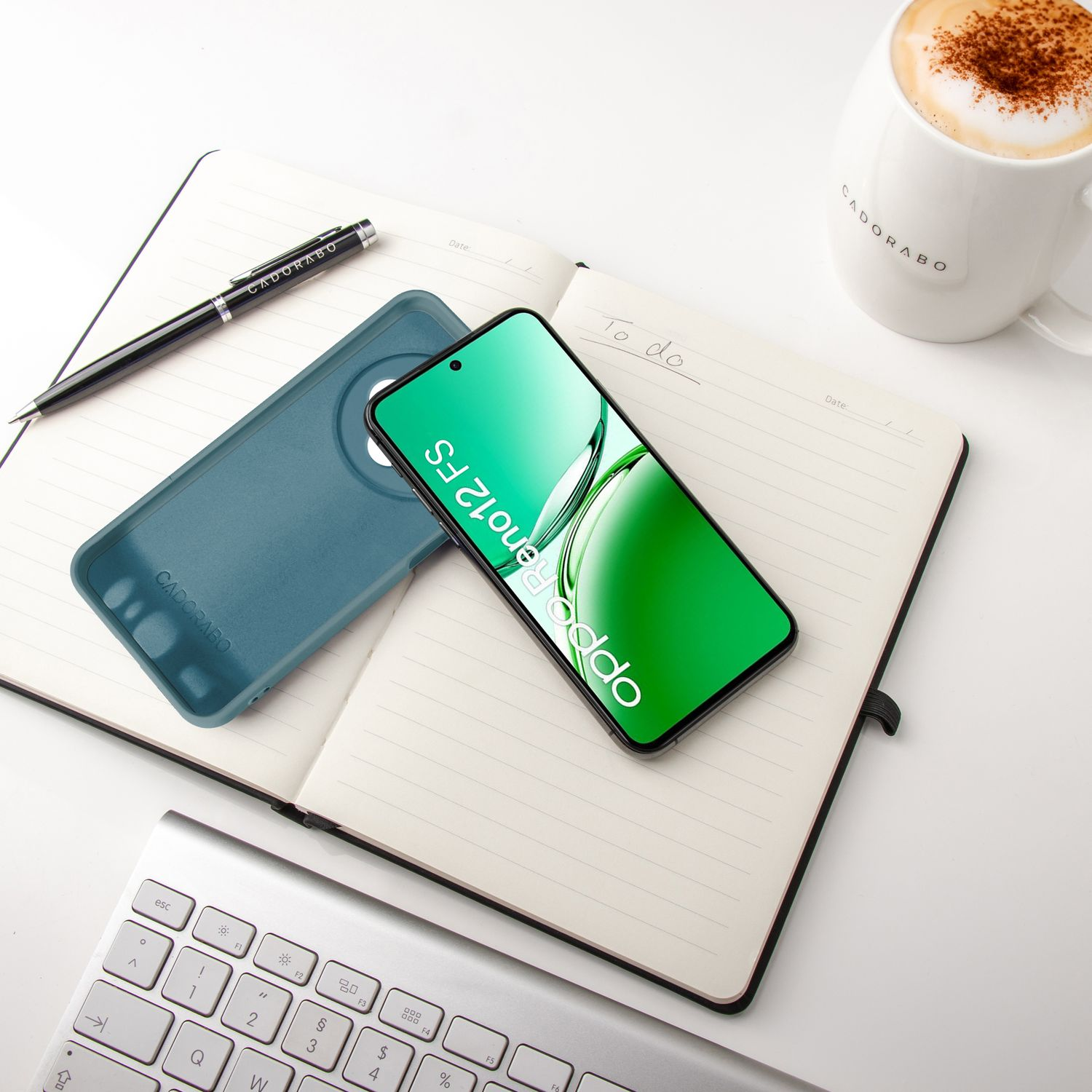 Ein Telefon, eine Hülle, ein Stift und eine Kaffeetasse liegen auf einem Notizbuch. Unten eine Tastatur.