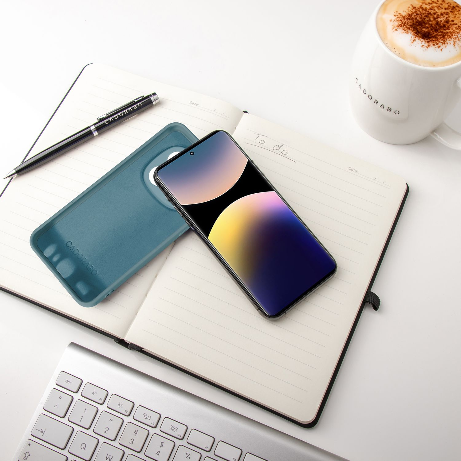 Telefon mit Hülle, Stift, Notizblock und Tasse. Die Szene ist auf einem weißen Schreibtisch.