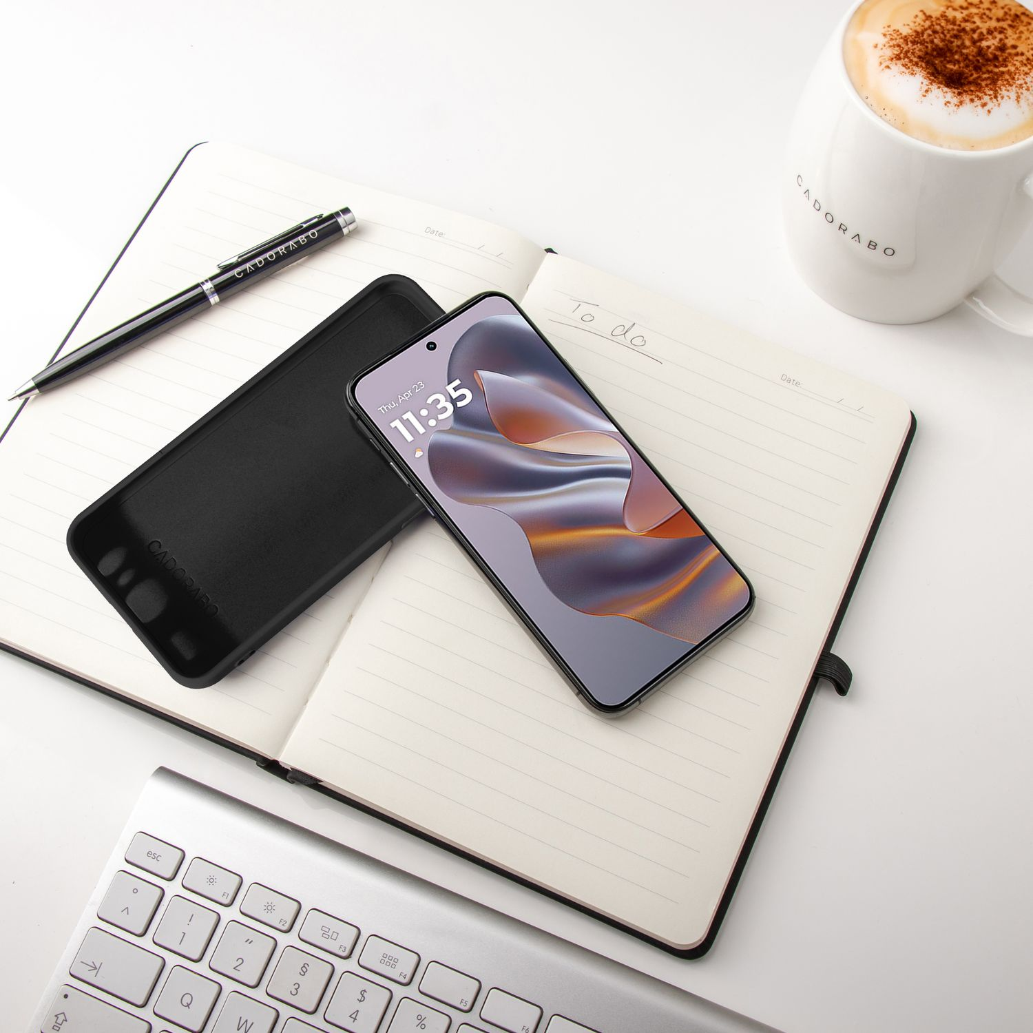 Telefon und schwarze Hülle auf einem Notizbuch mit Stift, eine Tasse Kaffee und Tastatur.