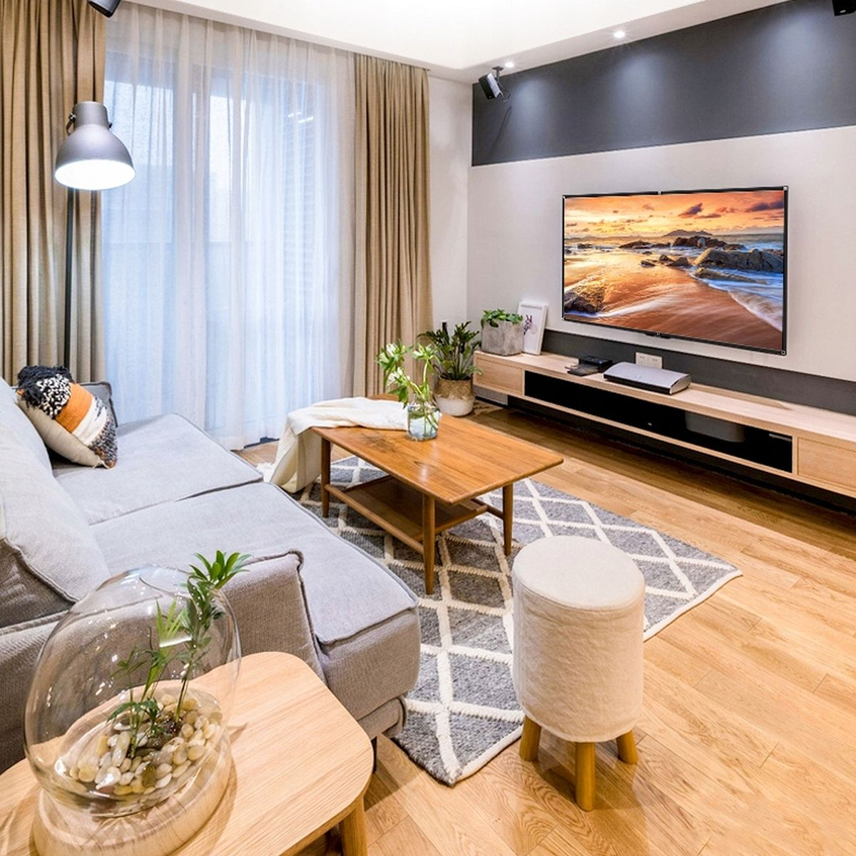 Salon z szarą sofą, drewnianym stołem i telewizorem. Zawiera rośliny, dywan i biały stołek.