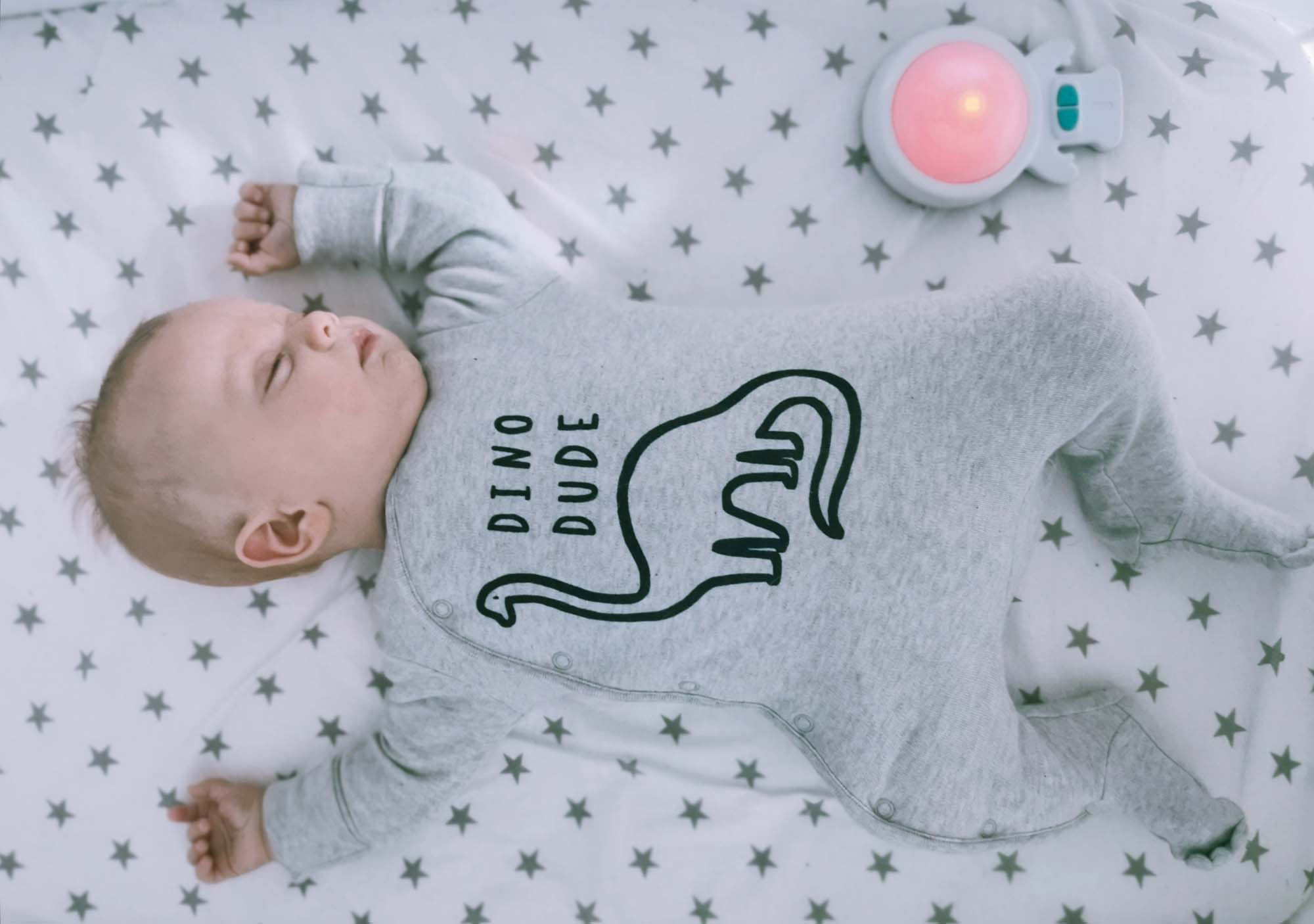 Baby schläft in einem Kinderbett und trägt einen grauen Strampler mit Dinosaurier-Grafik. Sterne auf dem Bettlaken.