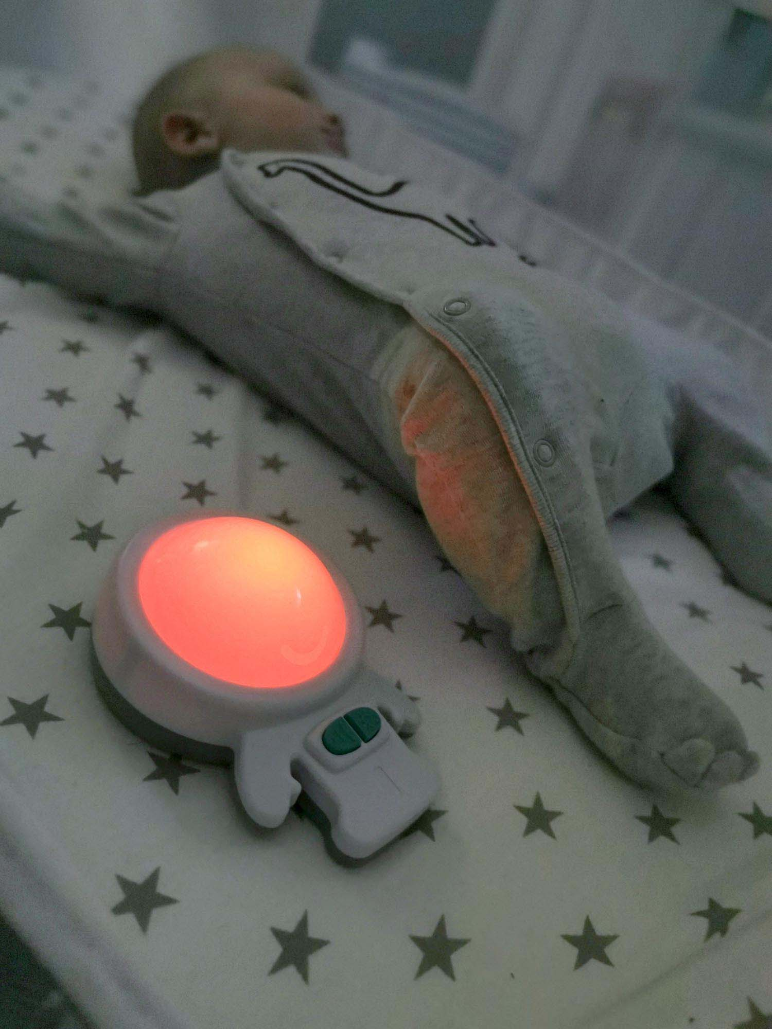 Ein Baby schläft in einem Kinderbett neben einem Nachtlicht. Das Nachtlicht ist weiß mit rotem Schein.