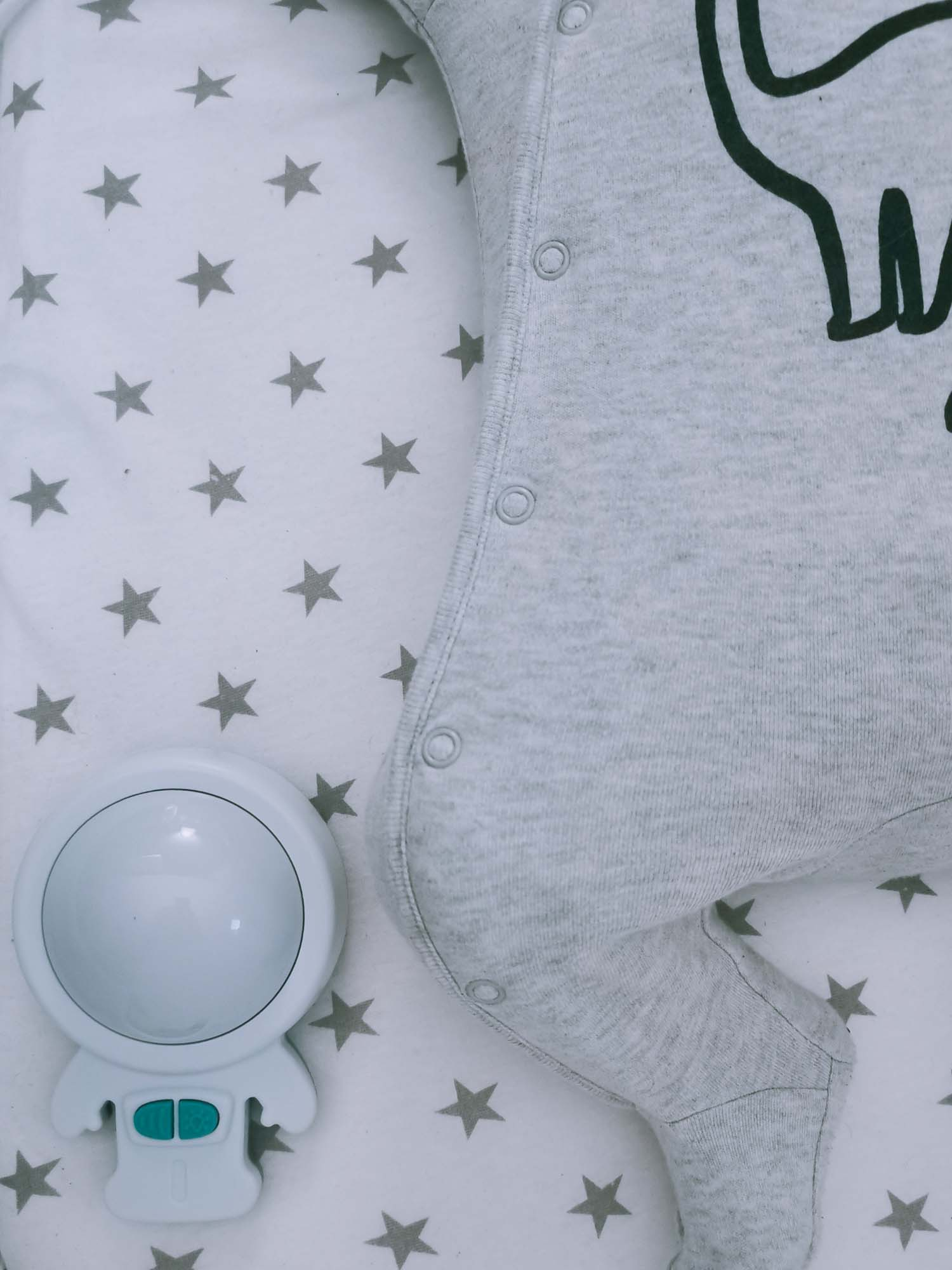 Baby in einem grauen Strampler auf einem Bett mit Sternenmuster. Ein Spielzeug in der Ecke.