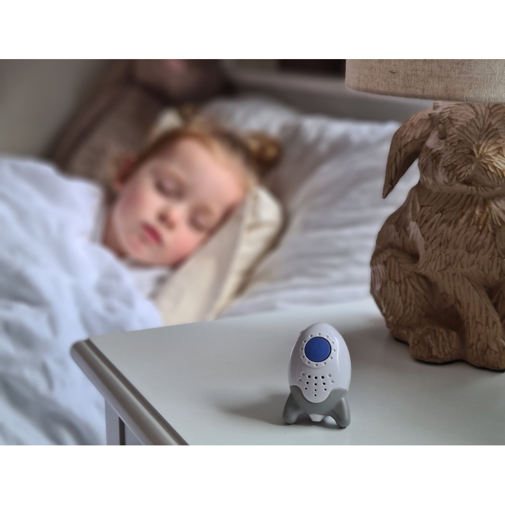 Ein Kind schläft in einem Bett. Ein raketenförmiges Geräuschgerät steht auf dem Nachttisch.