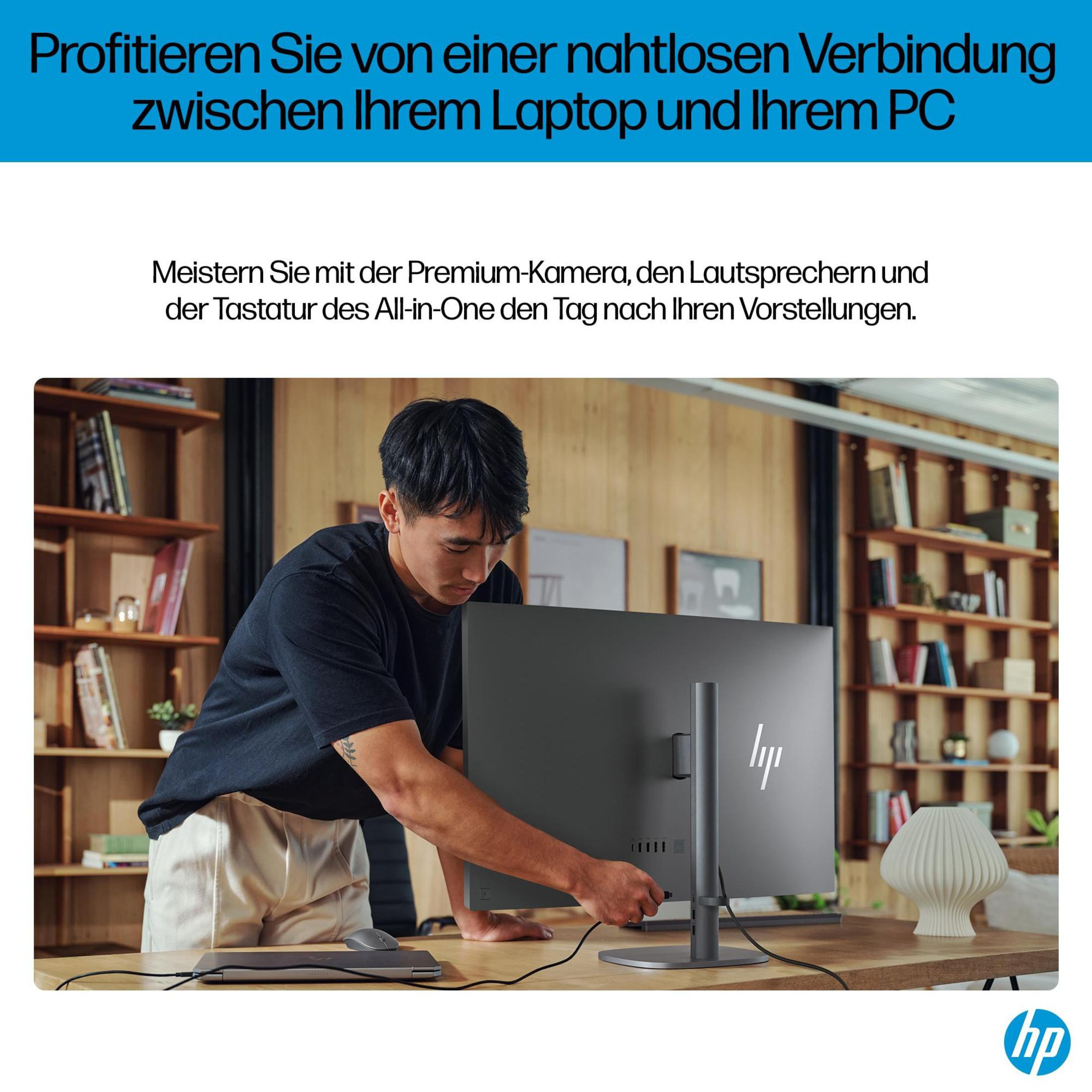 Ein Mann steckt ein Kabel in einen großen, grauen Monitor auf einem Schreibtisch. Im Hintergrund Fenster und ein Bücherregal.
