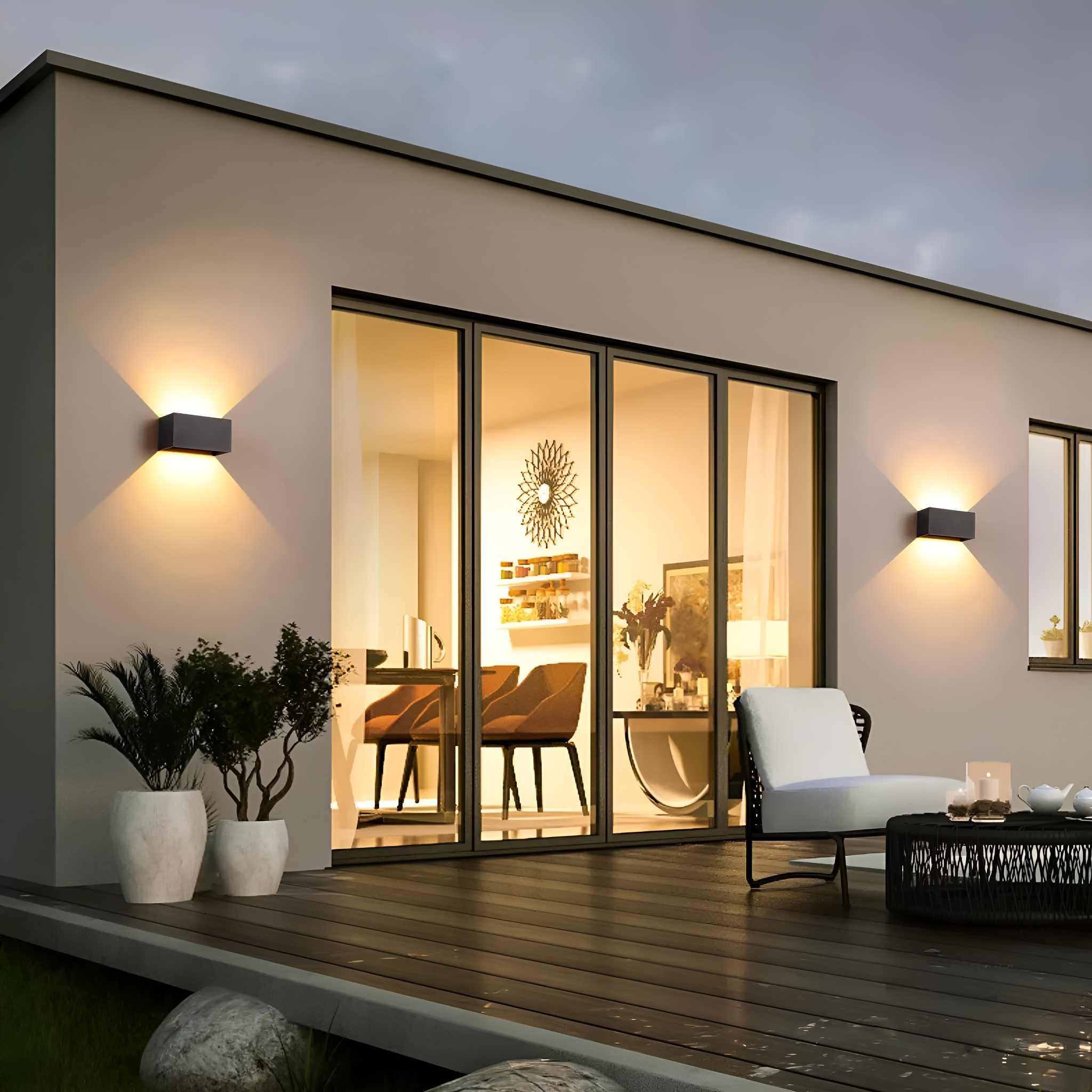 Hausfassade mit Lichtern, Holzterrasse und Glastüren mit Innenmöbeln.