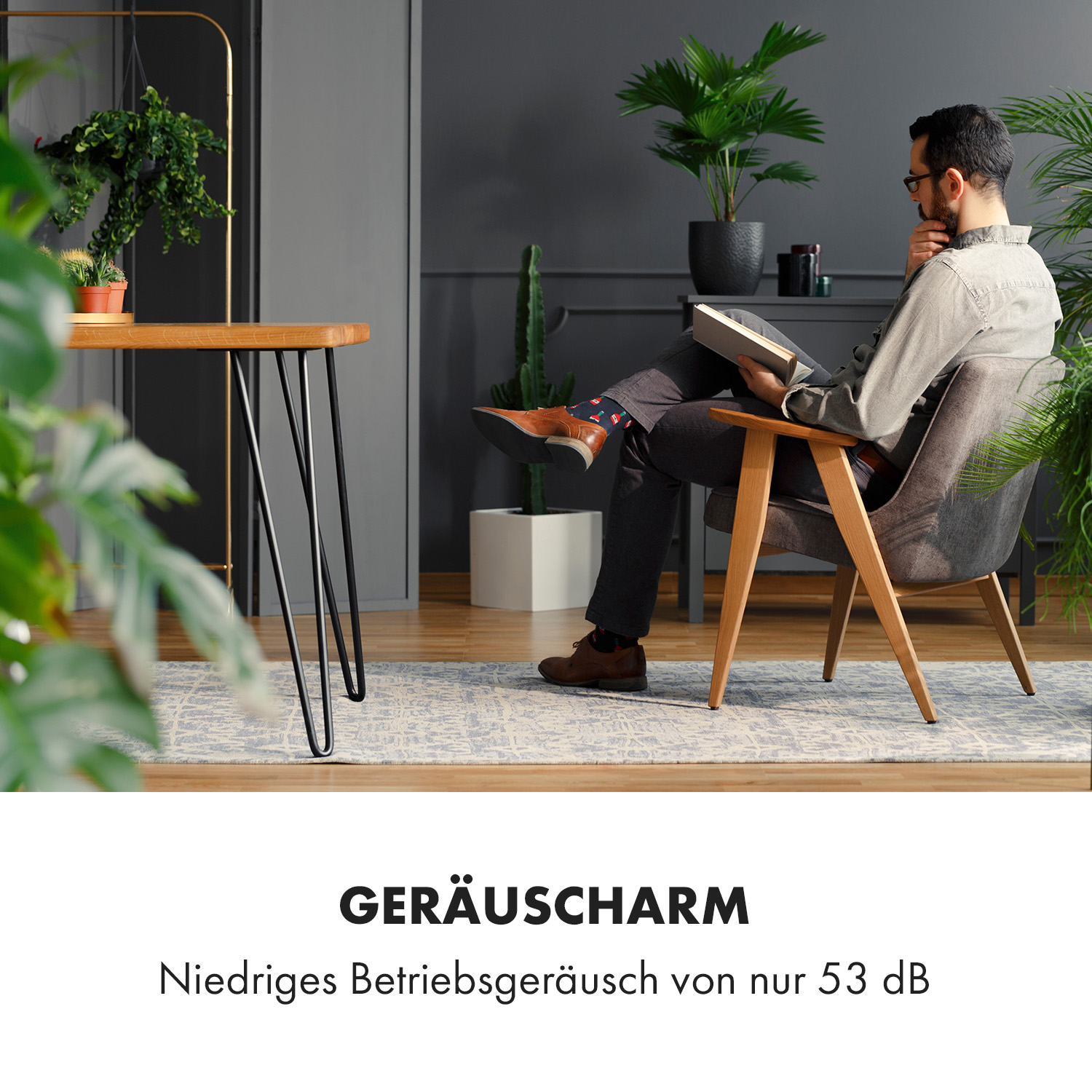 Ein Mann liest ein Buch in einem Raum. Ein Tisch mit Pflanze und ein grauer Stuhl. Pflanzen im Hintergrund.