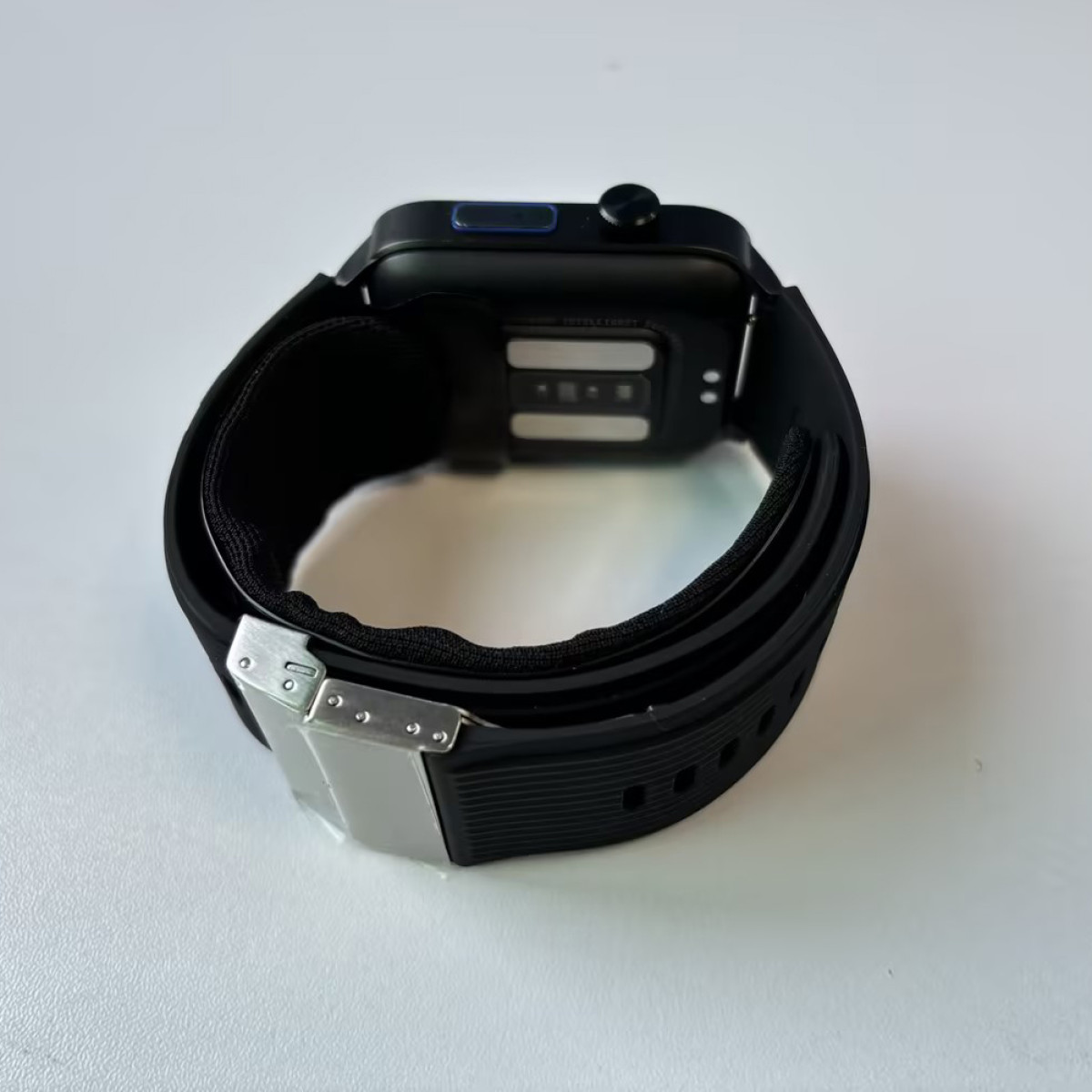 Czarny smartwatch z czarnym paskiem i srebrnym zapięciem, na białej powierzchni.