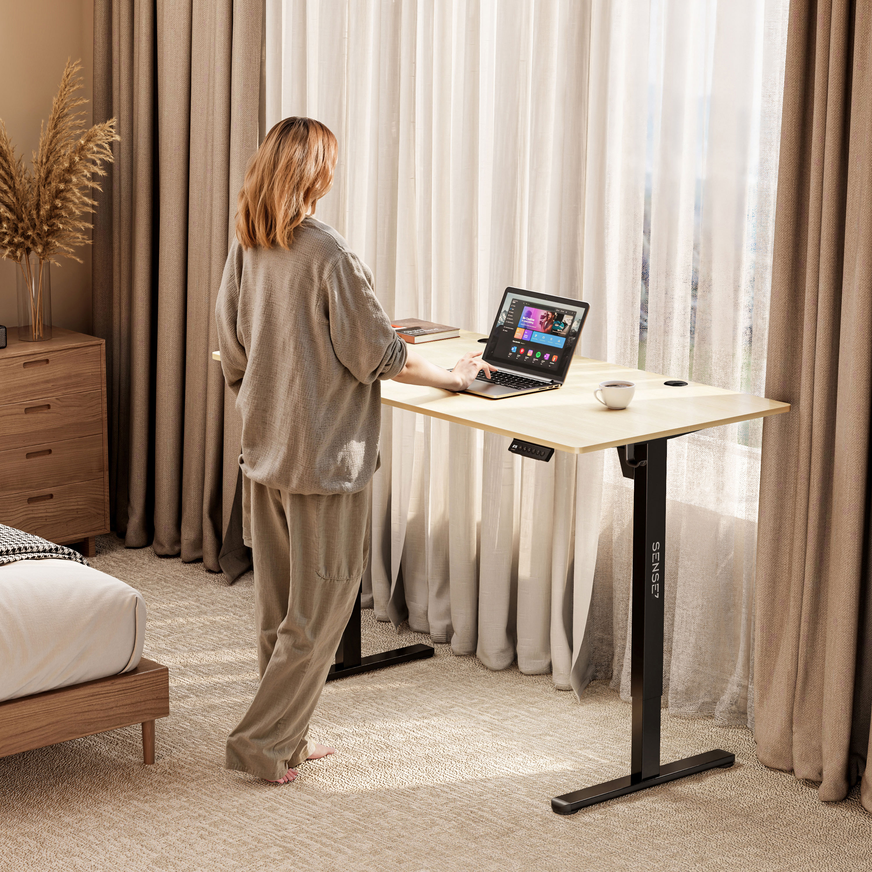 Vrouw die werkt aan een staande bureau bij een raam. Laptop open, koffiekopje en beige decoratie.