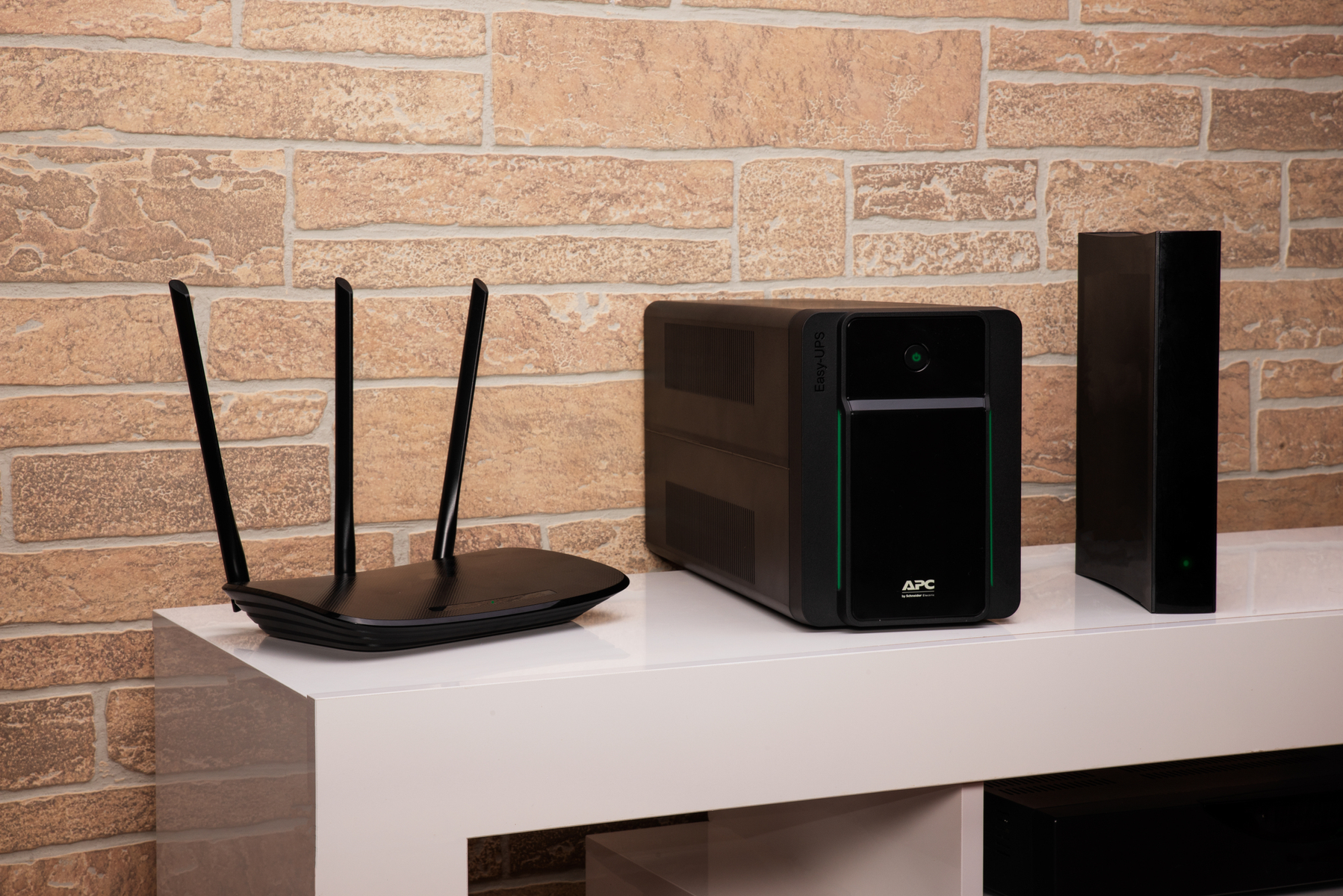 Ein Router, USV und ein kleines schwarzes Gerät auf einem weißen Regal vor einer Backsteinmauer.
