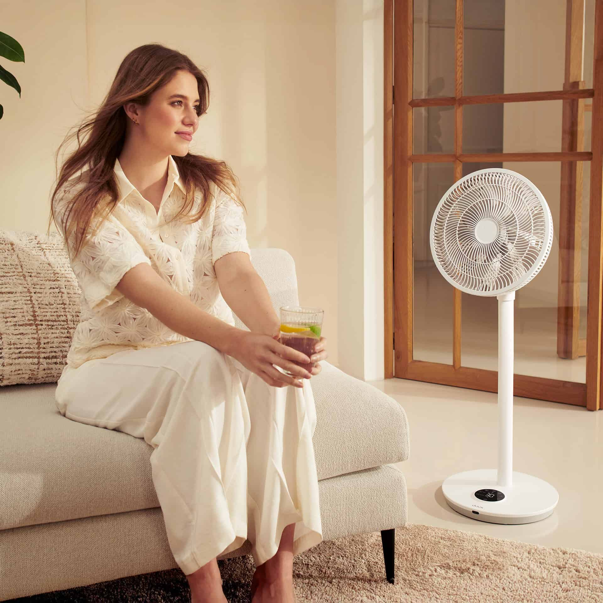 Frau sitzt auf Couch mit Ventilator und Glas Getränk.