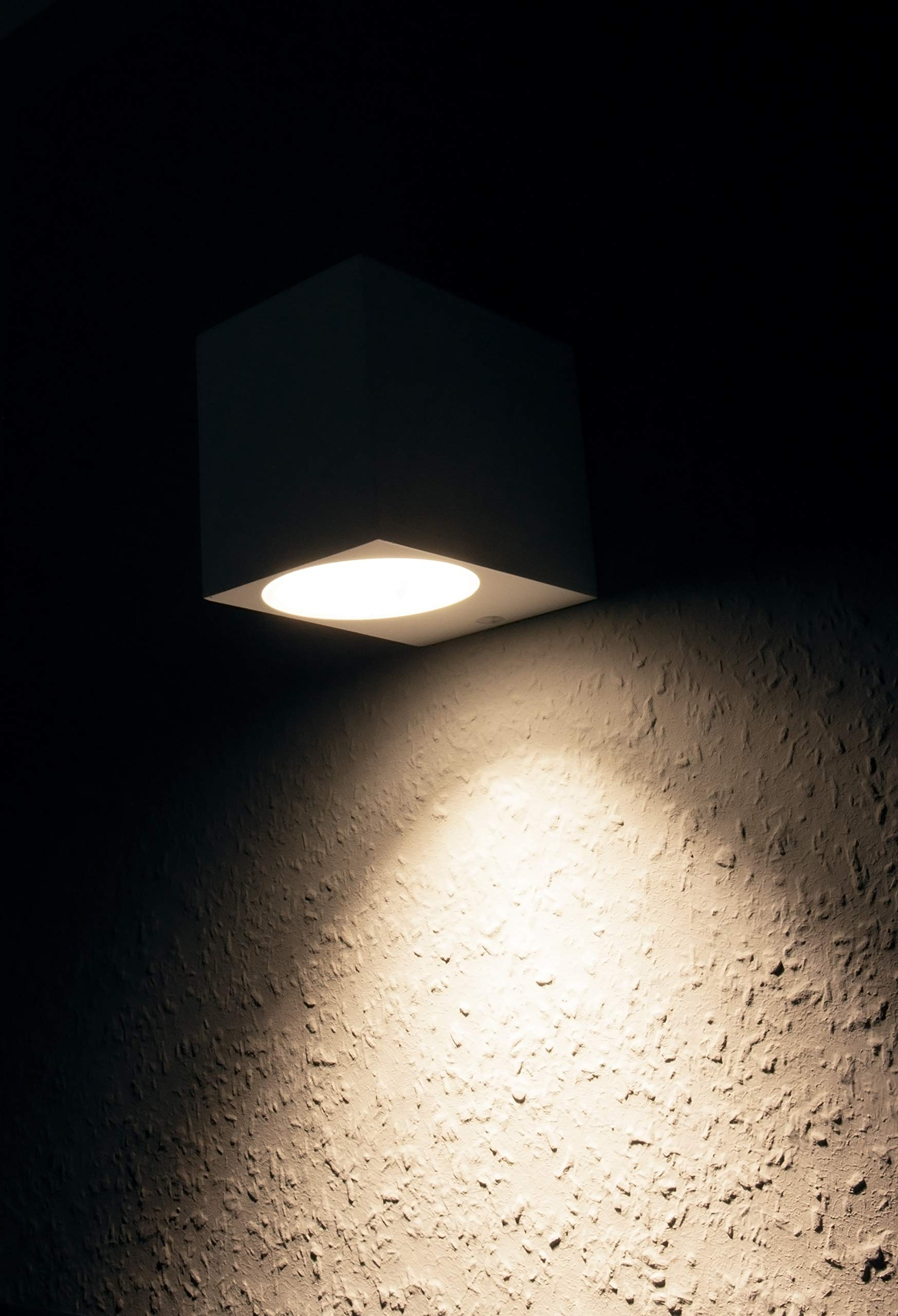 Eine quadratische weiße Deckenleuchte ist an einer strukturierten, dunkelgrauen Wand montiert und wirft einen hellen Lichtkreis.