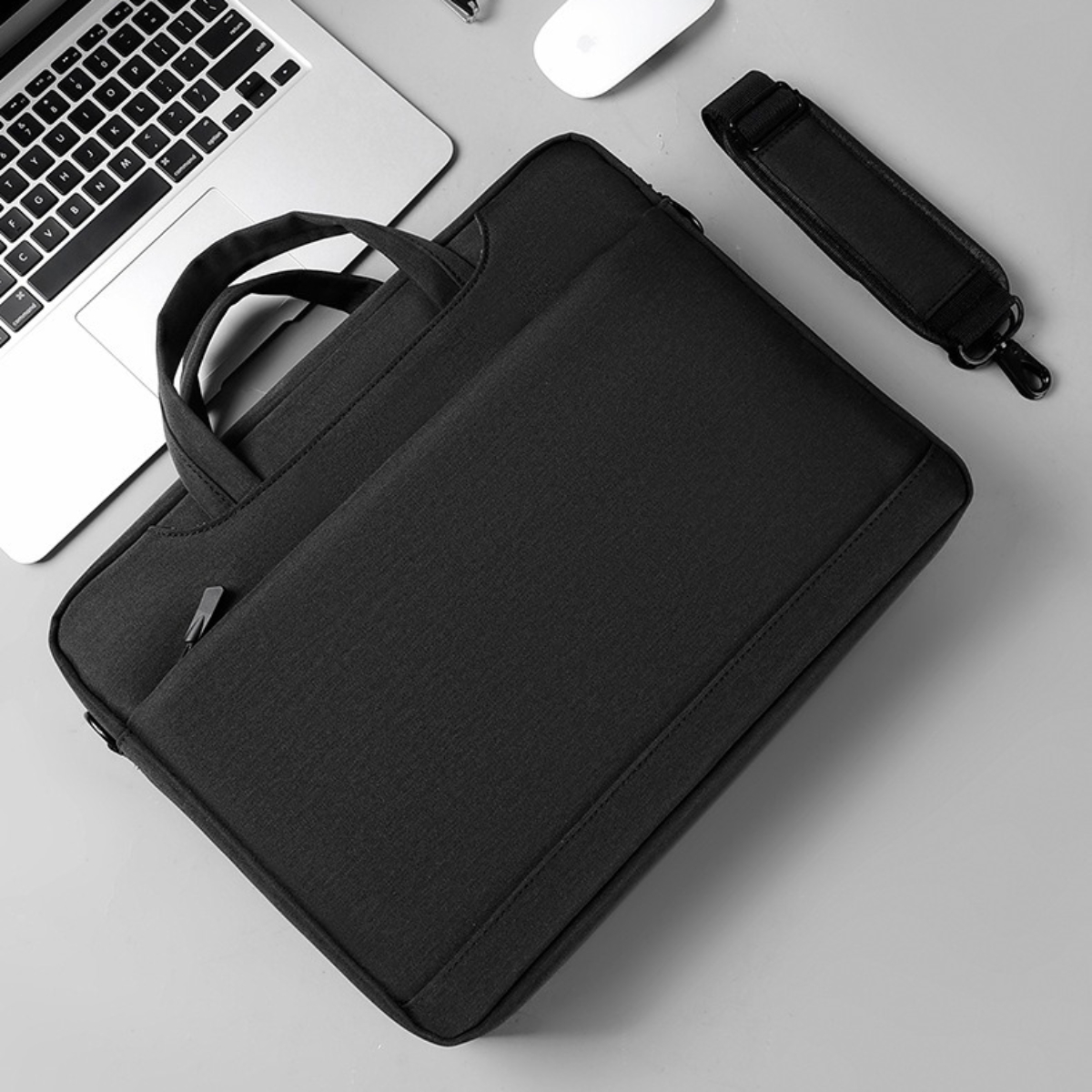 Schwarze Laptoptasche mit Griff und Schultergurt auf grauer Oberfläche. Weiße Maus und Laptop sichtbar.