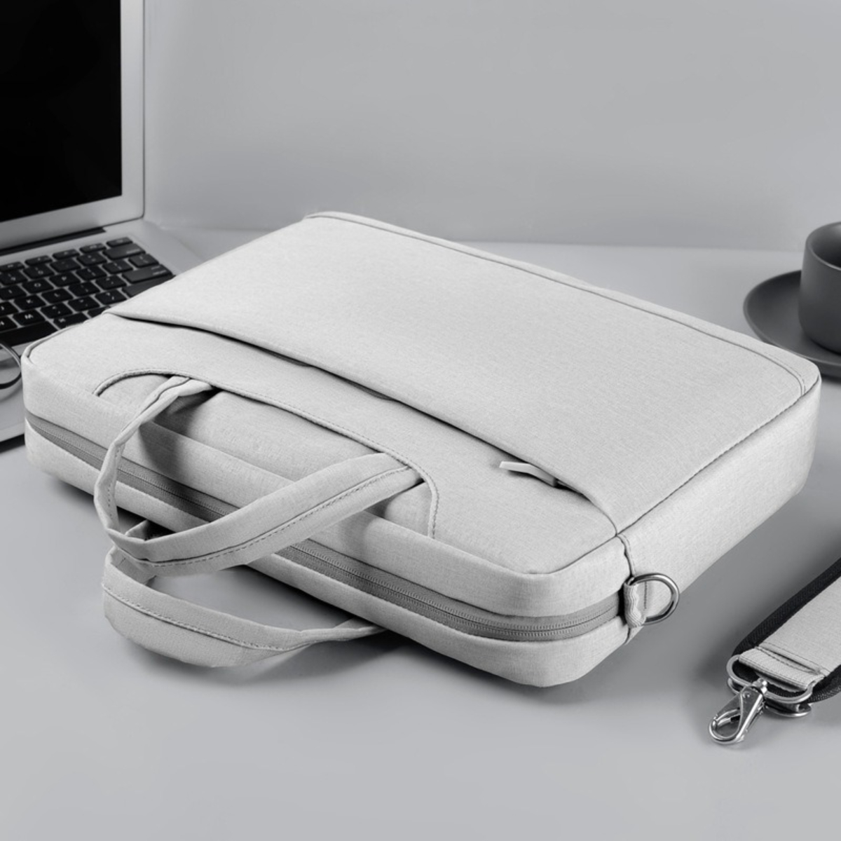 Hellgraue Laptoptasche mit Griff und Schultergurt. Ein Laptop steht auf einem Schreibtisch.
