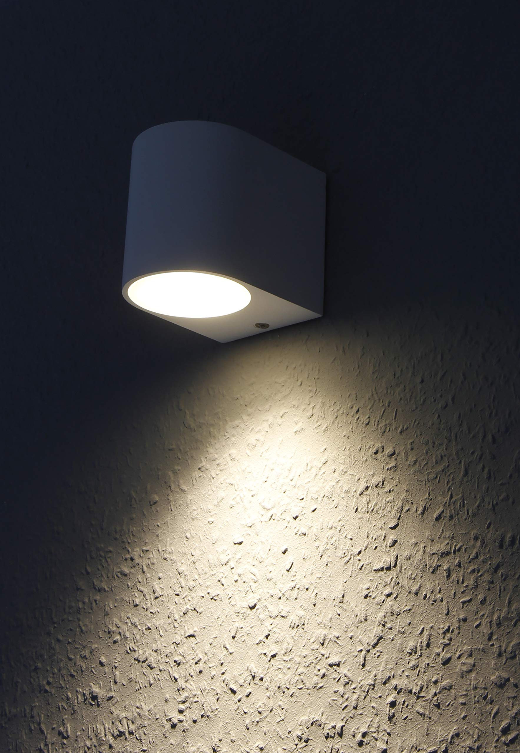 Weiße, ovale Außenwandleuchte. Das Licht scheint nach unten auf eine strukturierte Wand. Der Hintergrund ist dunkel.