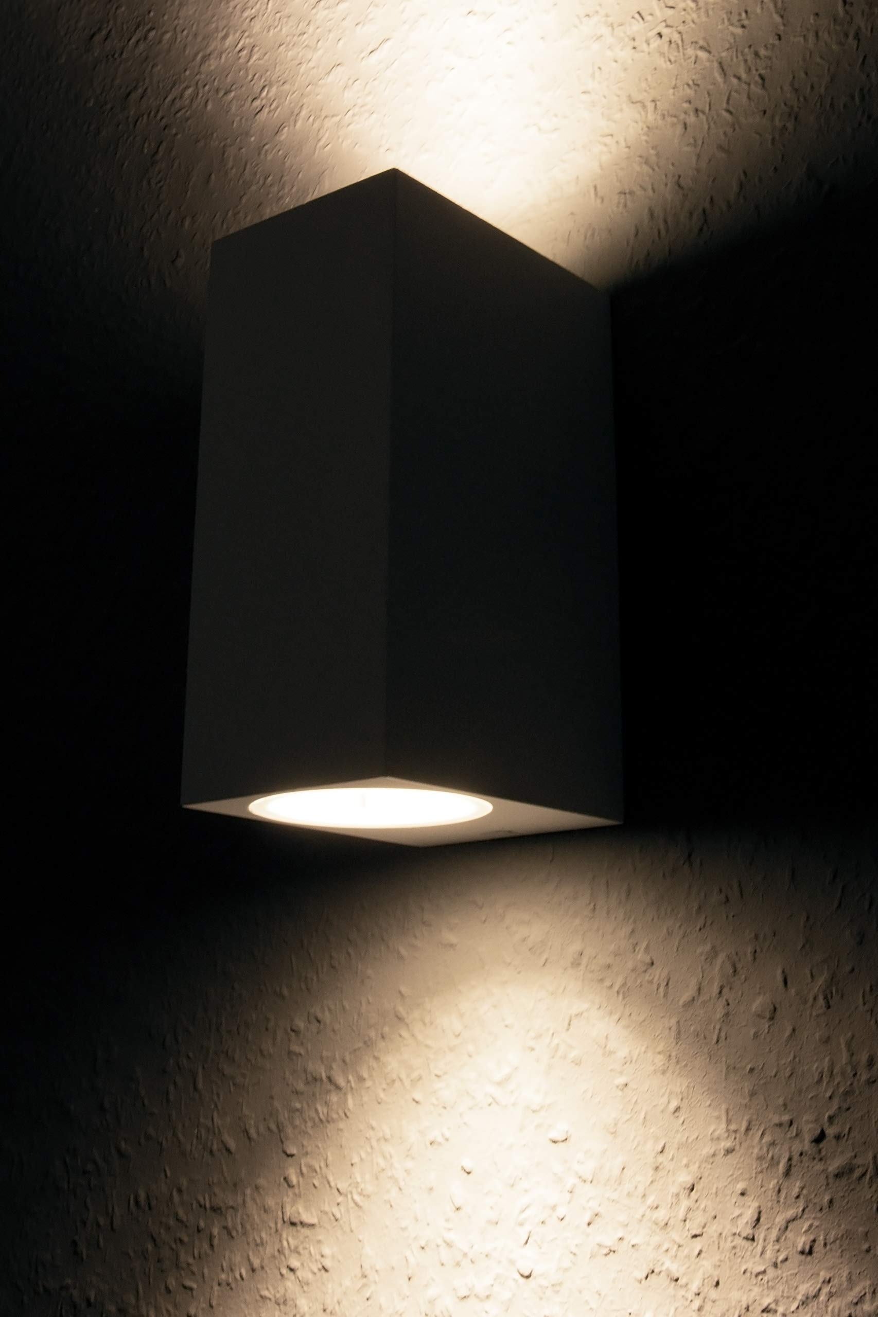 Wandmontierte weiße Leuchte strahlt Licht aus, vor dunklem Hintergrund.
