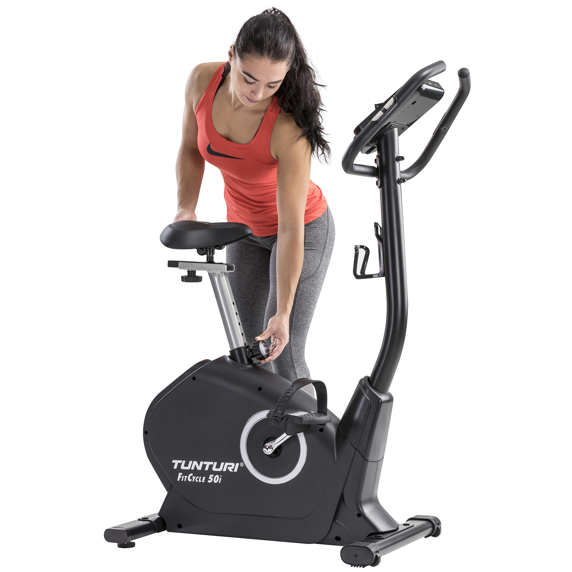 Vrouw past hometrainer aan. Rode top, grijze broek. Zwarte fiets met display.