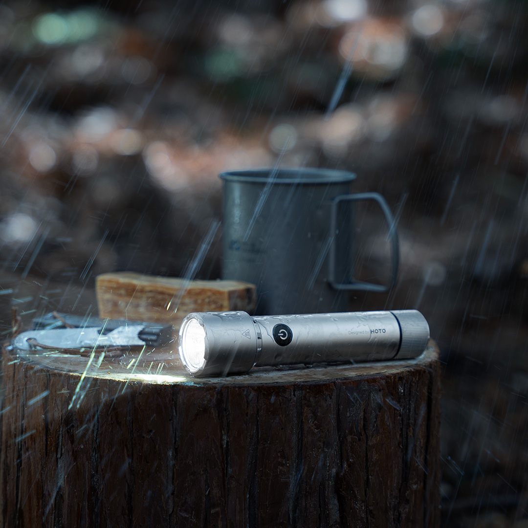 Silberne Taschenlampe auf einem Baumstumpf im Regen, mit Messer und Becher in der Nähe.