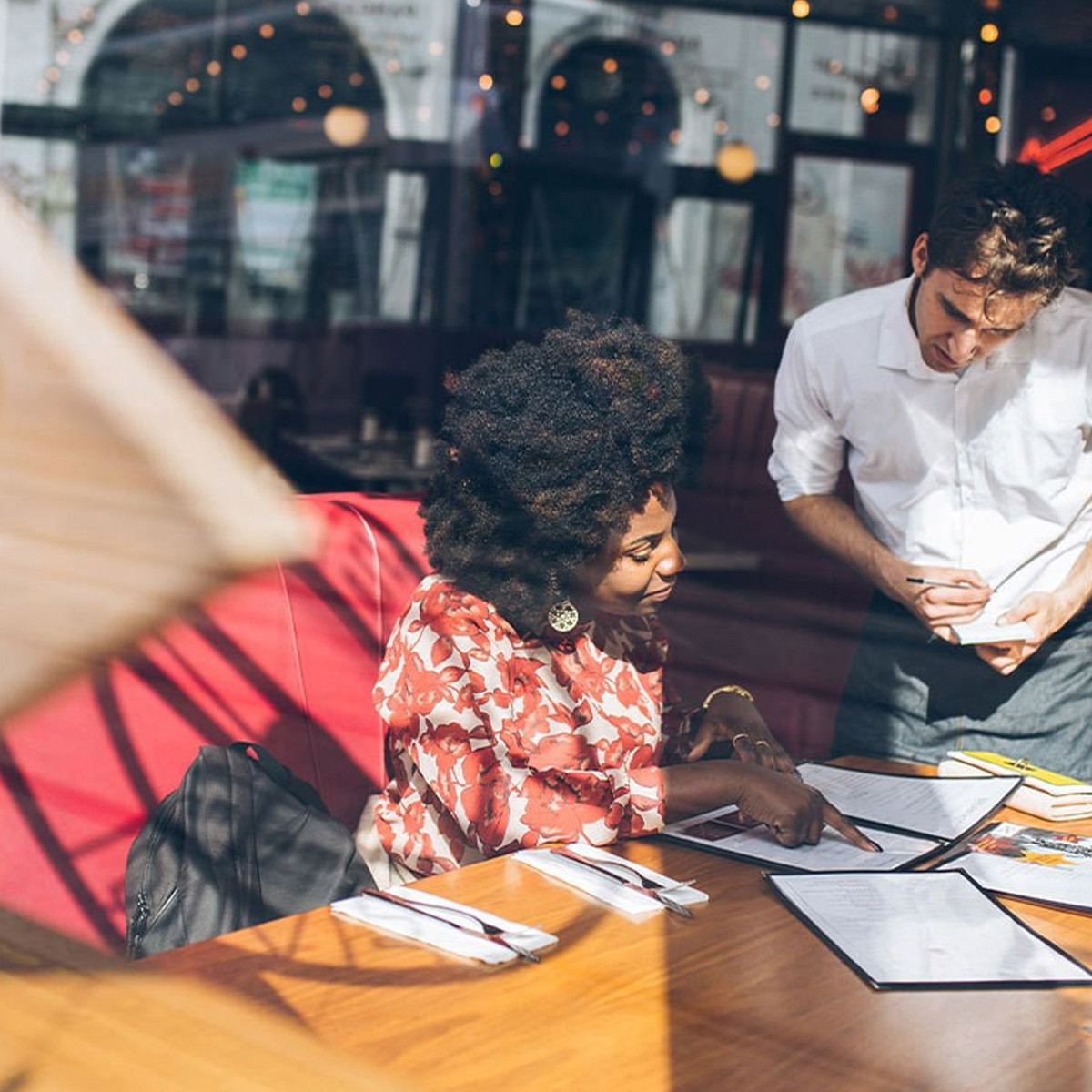 Restaurantszene mit einer Frau, die auf eine Speisekarte zeigt, und einem Kellner, der Notizen macht, in hellem Licht.