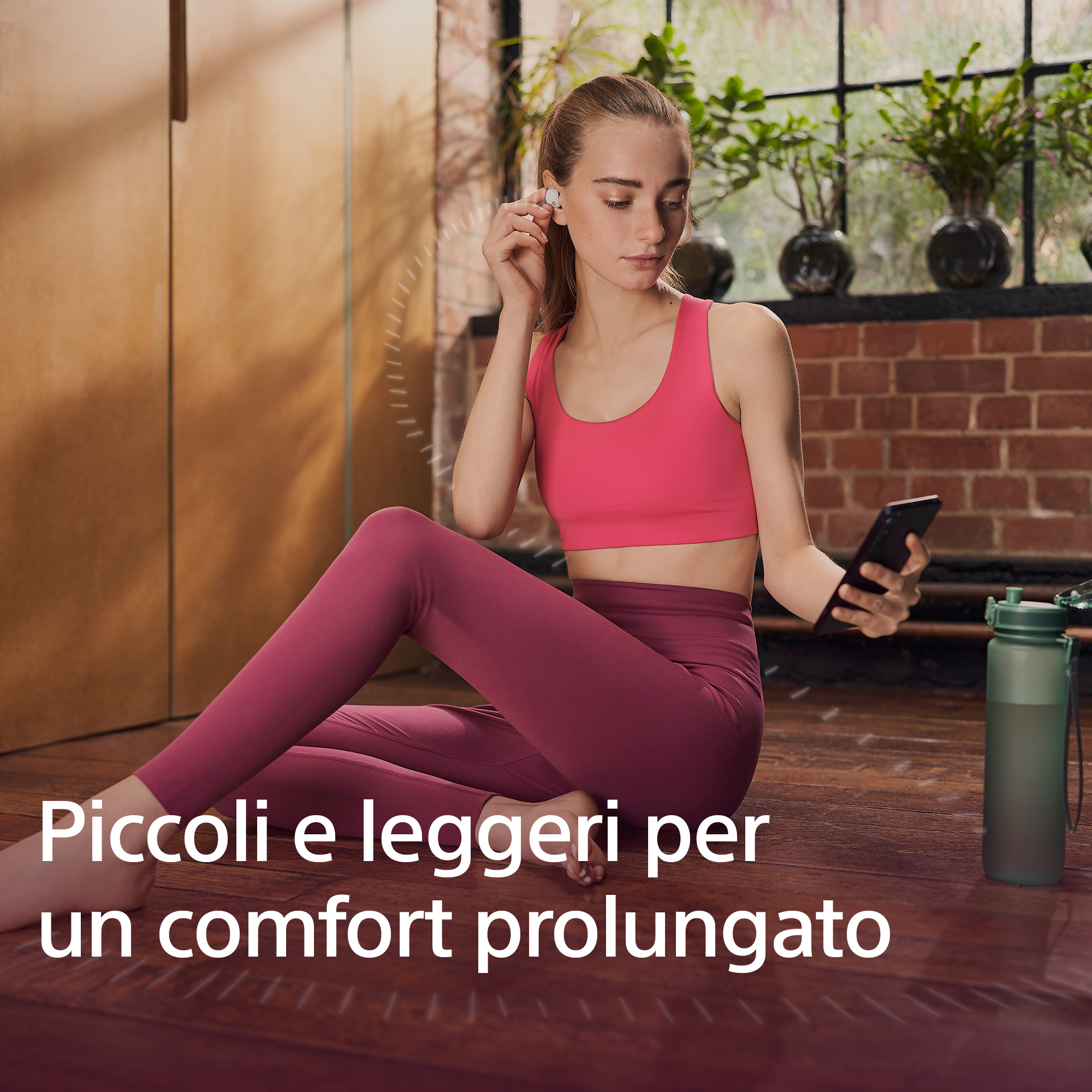 Mujer sosteniendo un teléfono, usando top y leggings rosas, usando auriculares. Botella de agua en el suelo.