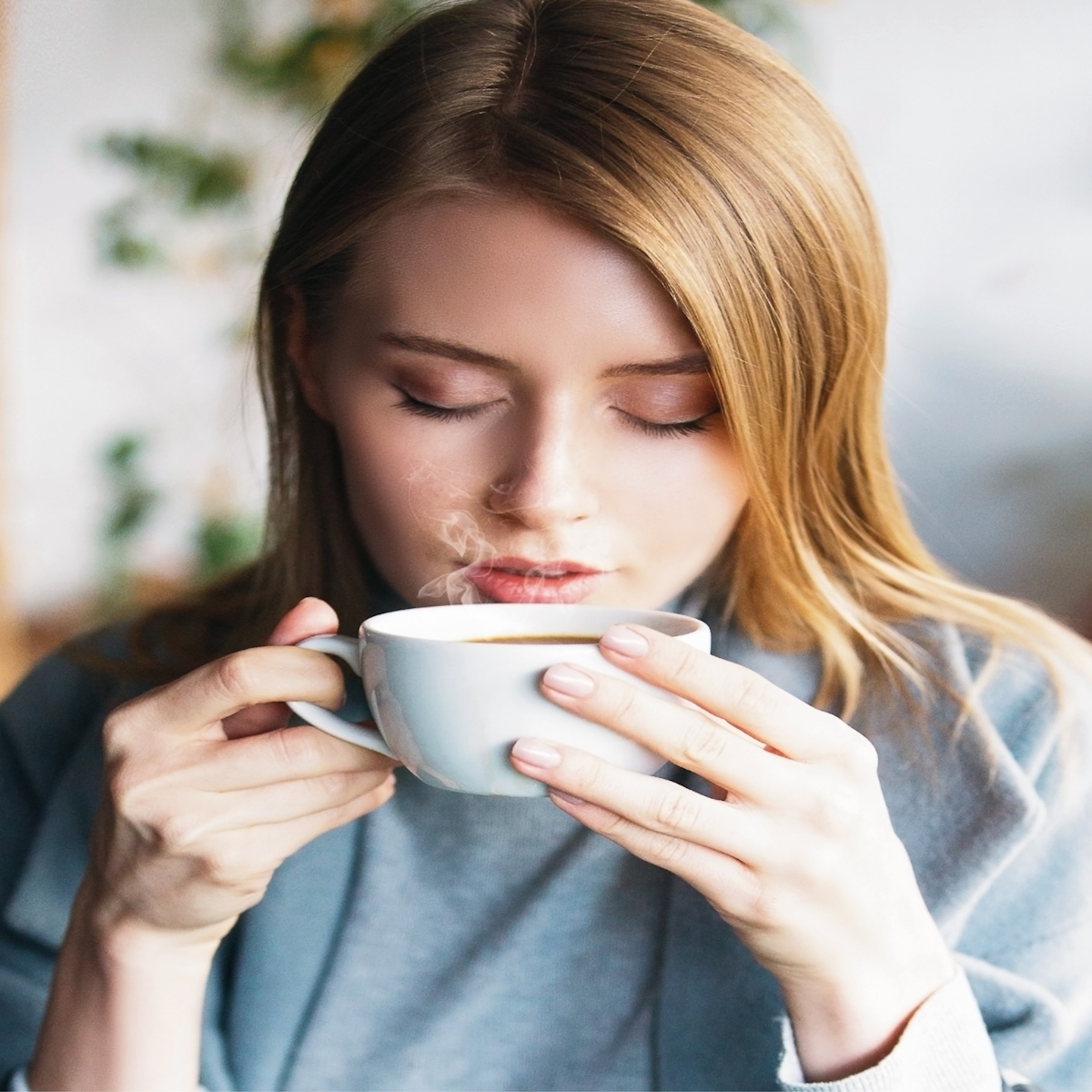 Frau hält weiße Tasse, trinkt, Augen geschlossen, Dampf sichtbar.