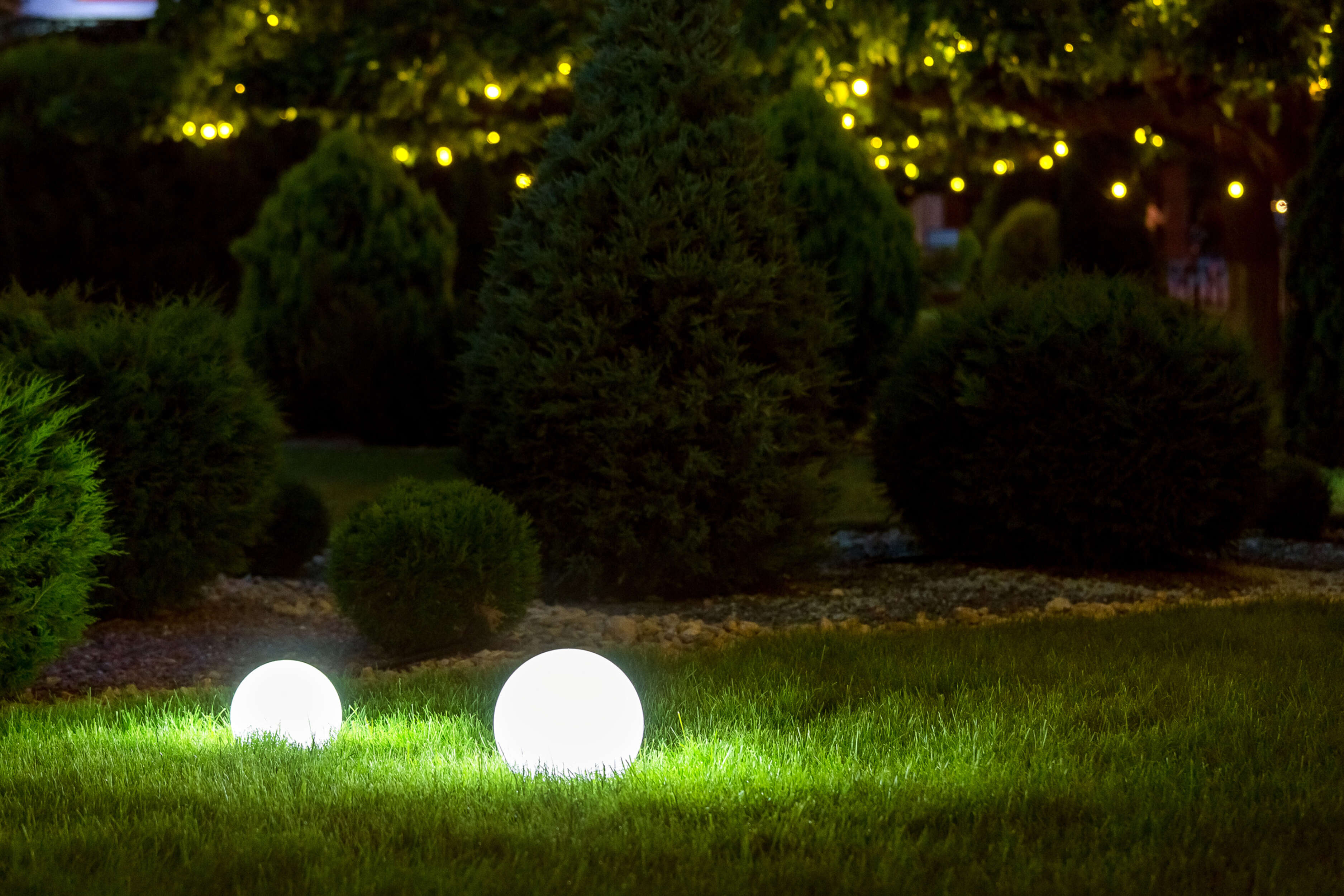 Zwei leuchtende weiße Kugeln auf grünem Gras bei Nacht. Dunkler Hintergrund mit Bäumen und Lichterketten.