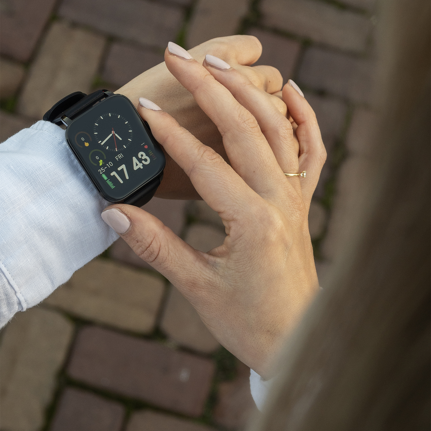 De hand van een persoon draagt een zwarte smartwatch, die de tijd en datum weergeeft, op een bakstenen oppervlak.