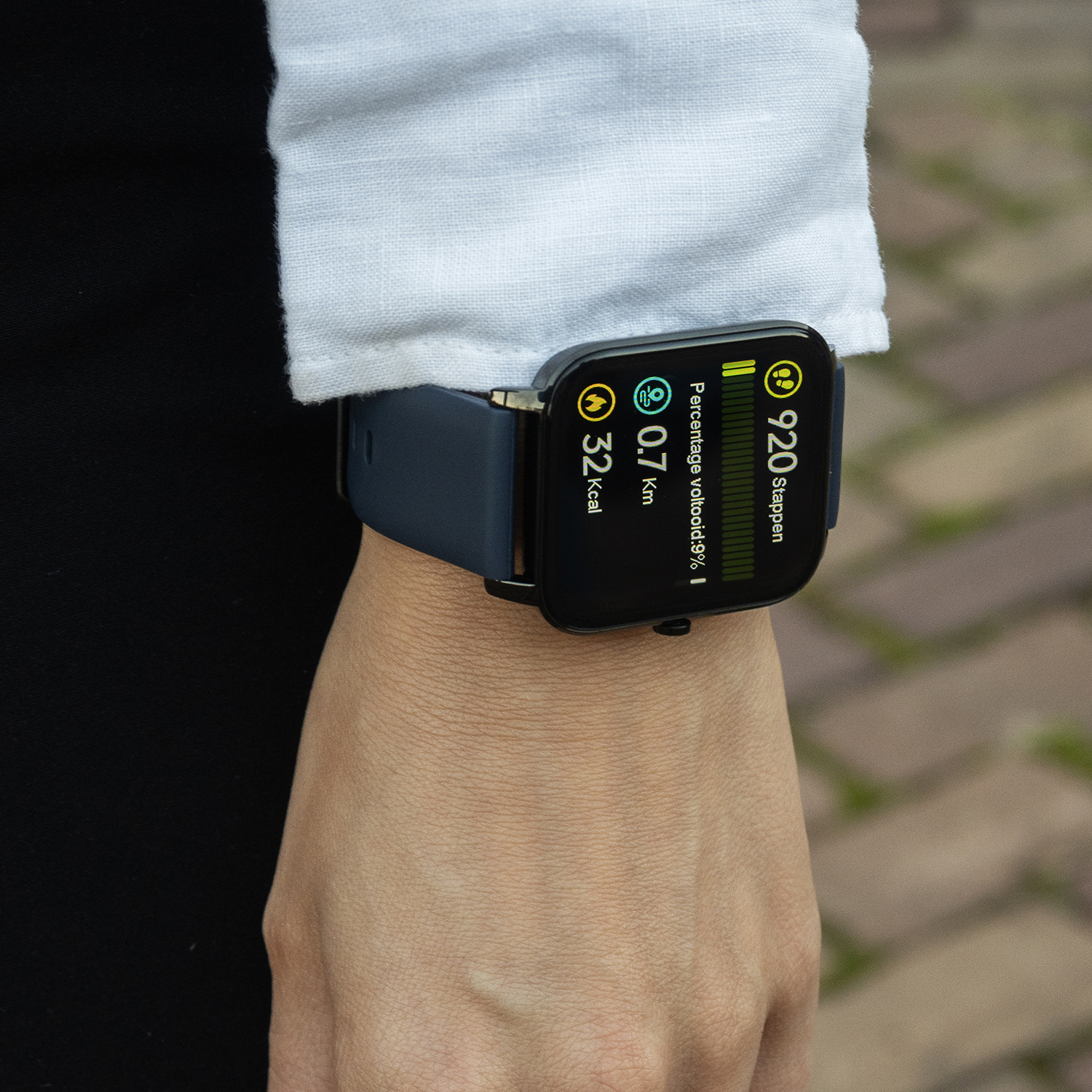 Close-up van een smartwatch op een pols. Scherm toont activiteitengegevens. De persoon draagt een wit shirt.