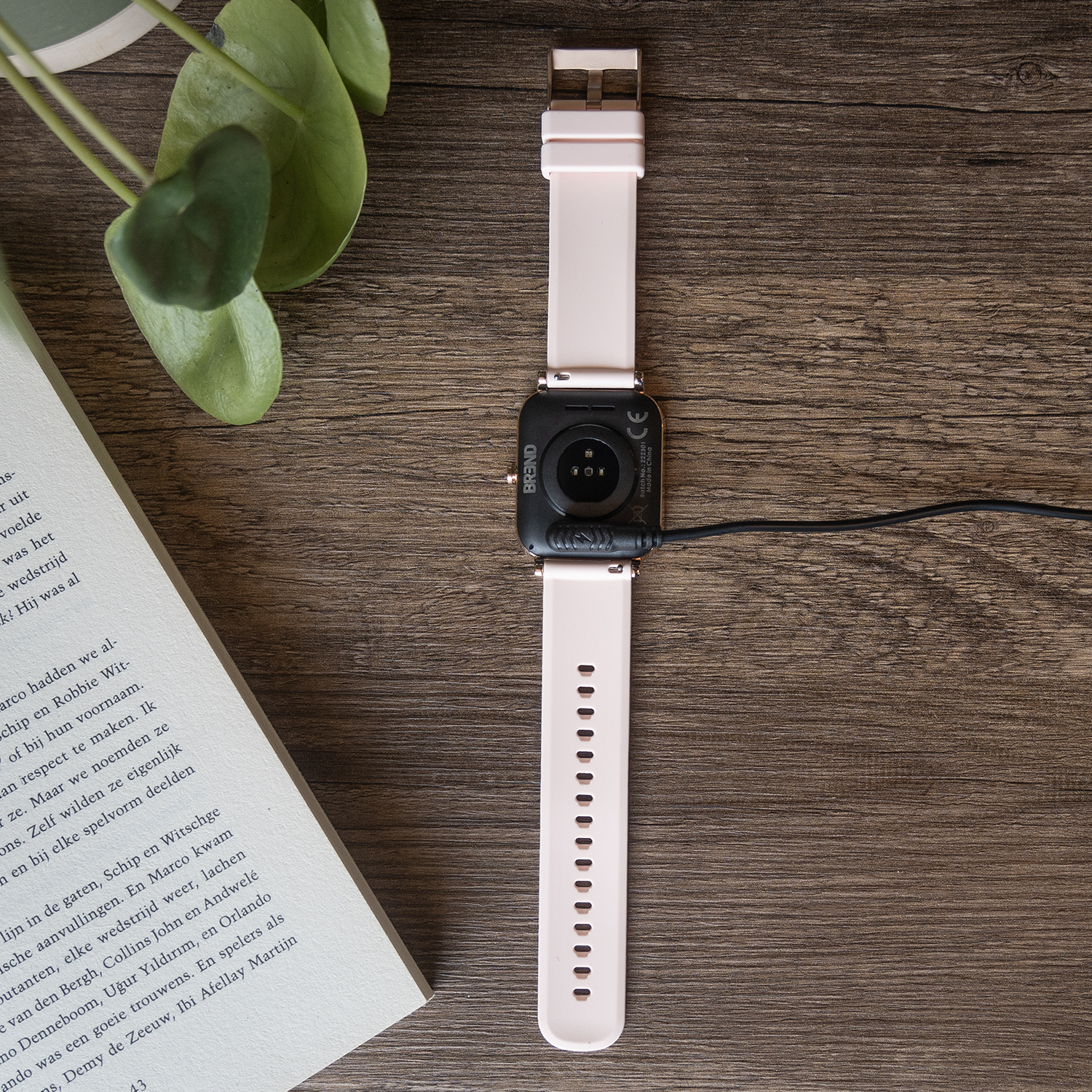 Smartwatch met roze bandje aangesloten op een oplader. Ligt op een houten tafel.