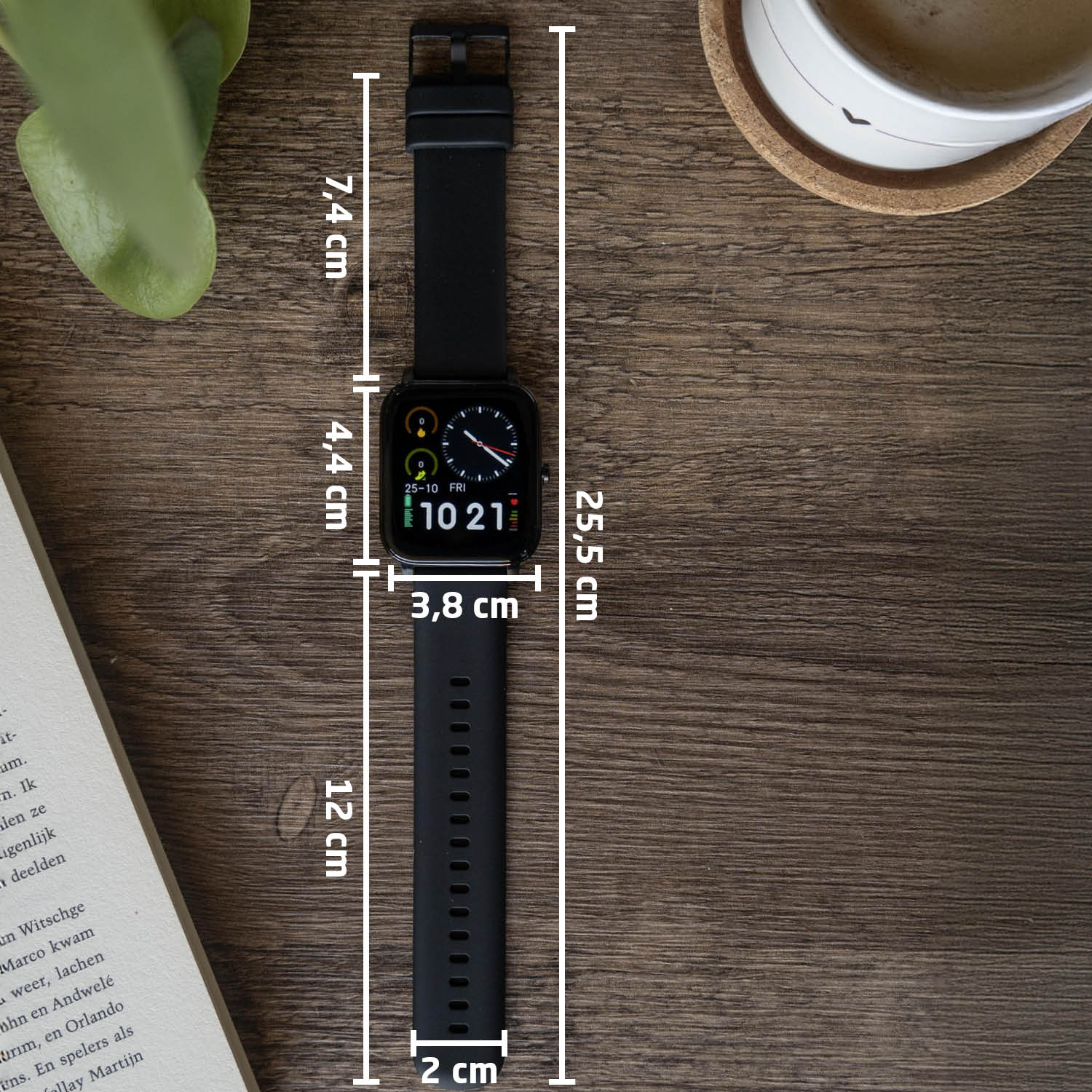 Een zwarte smartwatch met een zwarte band wordt met afmetingen weergegeven op een houten oppervlak. Er is een boek en een plant.