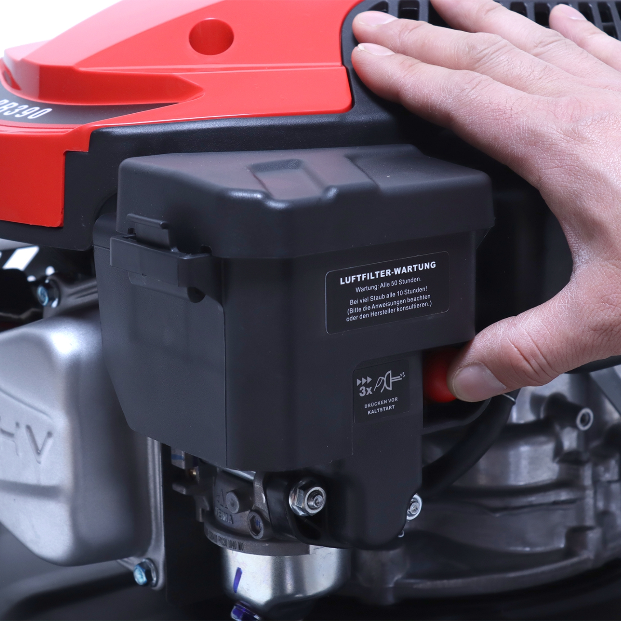 Eine Hand auf einer rot-schwarzen Motorabdeckung mit Text zur Wartung. Ein roter Startknopf ist sichtbar.