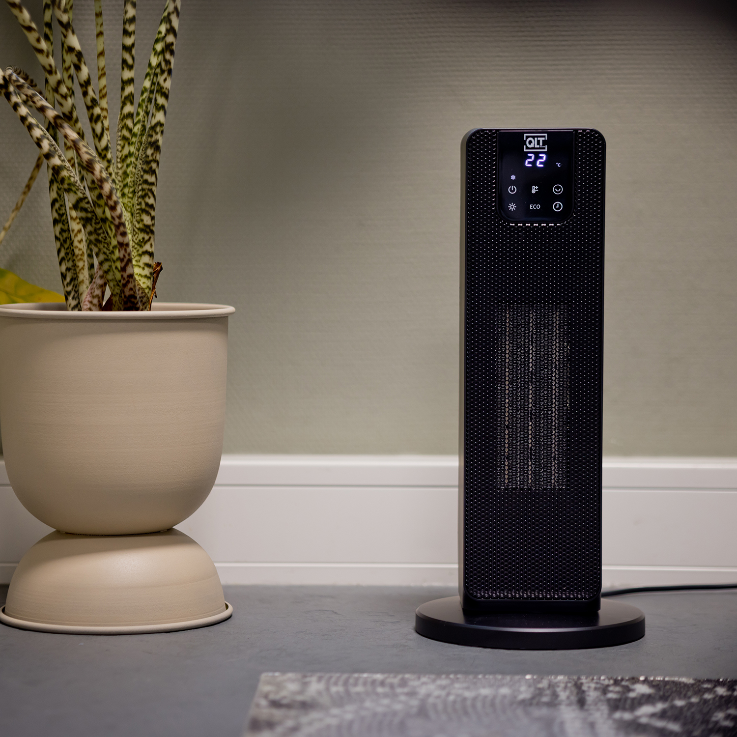 Black tower heater with a digital display, next to a plant in a beige pot. The heater is on a dark base.