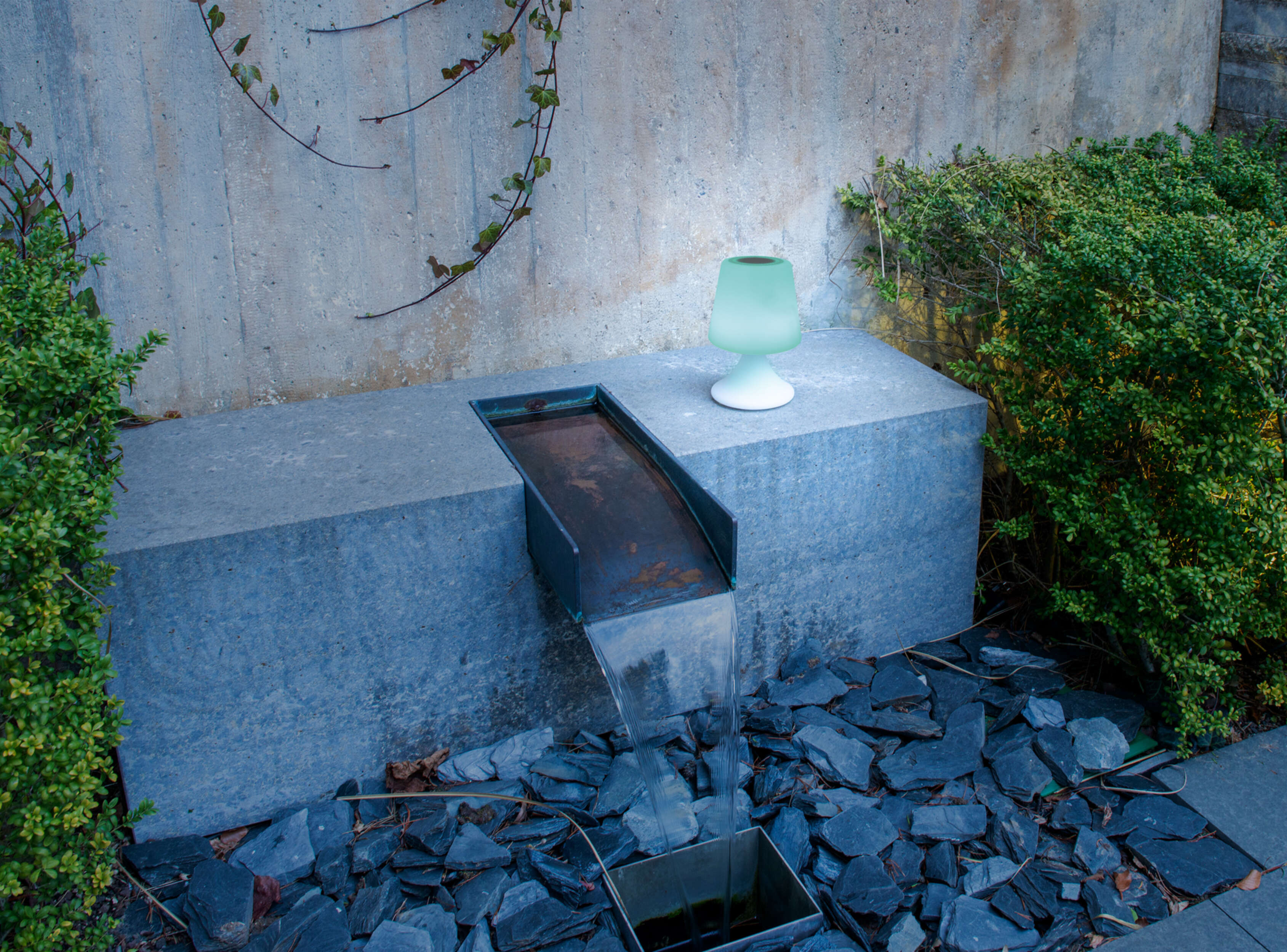 Wasserspiel mit kleiner grüner Lampe auf einer Steinbank. Wasser fließt in ein Becken.