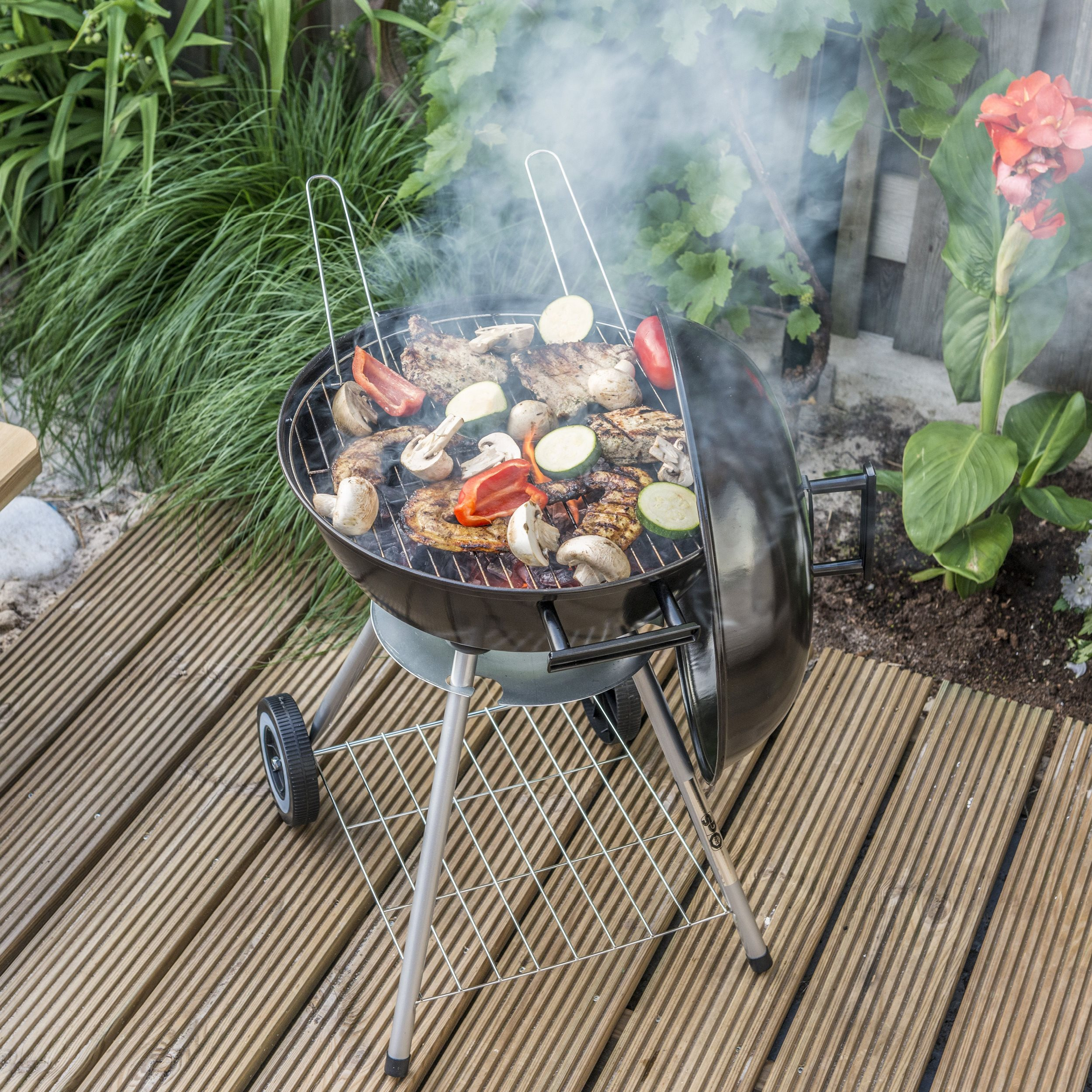Een open grill met voedsel dat gaart en rook. Grill op een houten terras met planten.