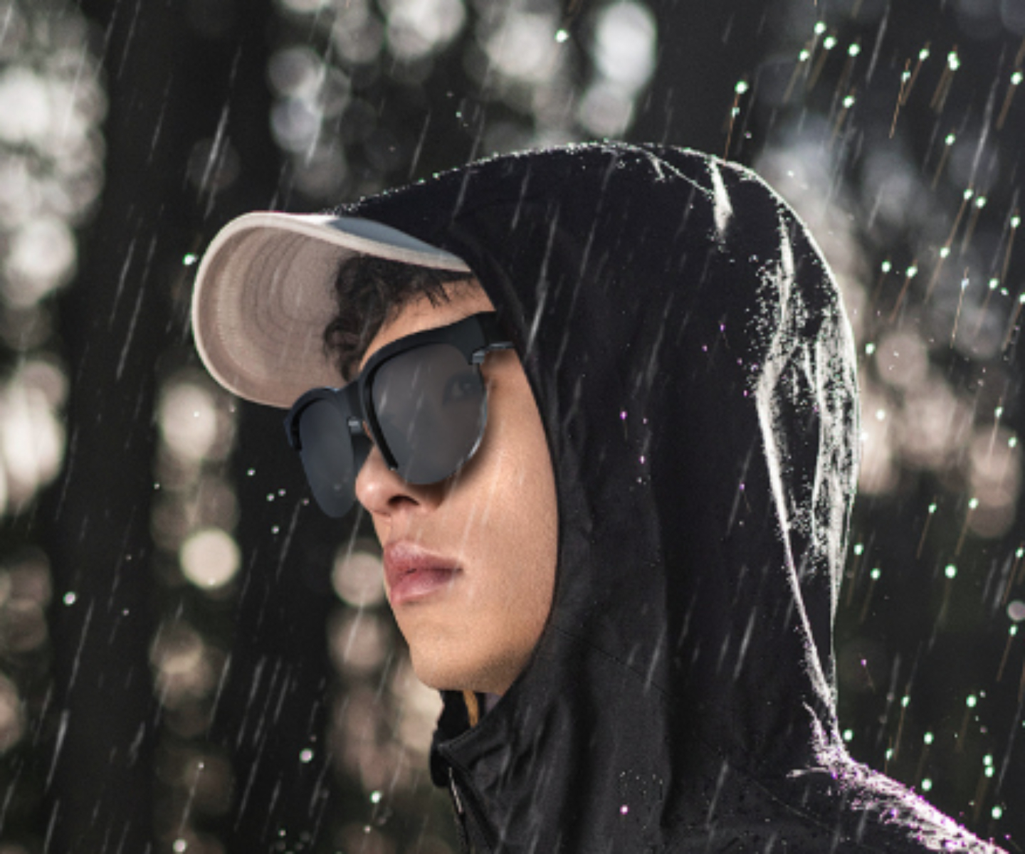 Ein Mann mit Sonnenbrille und Kapuze steht im Regen. Es regnet. Der Hintergrund ist dunkel.