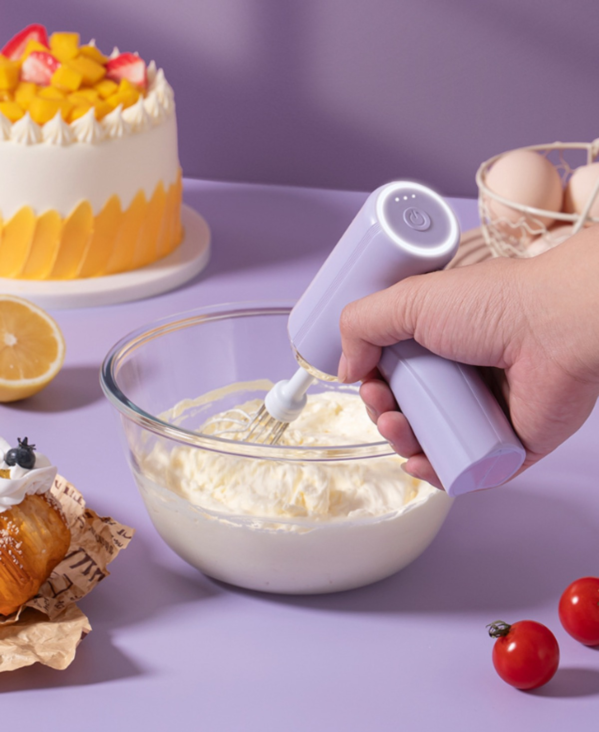 Die Hand einer Person verwendet einen lavendelfarbenen elektrischen Mixer, um Sahne in einer Glasschüssel zu schlagen. Ein Kuchen ist im Hintergrund.