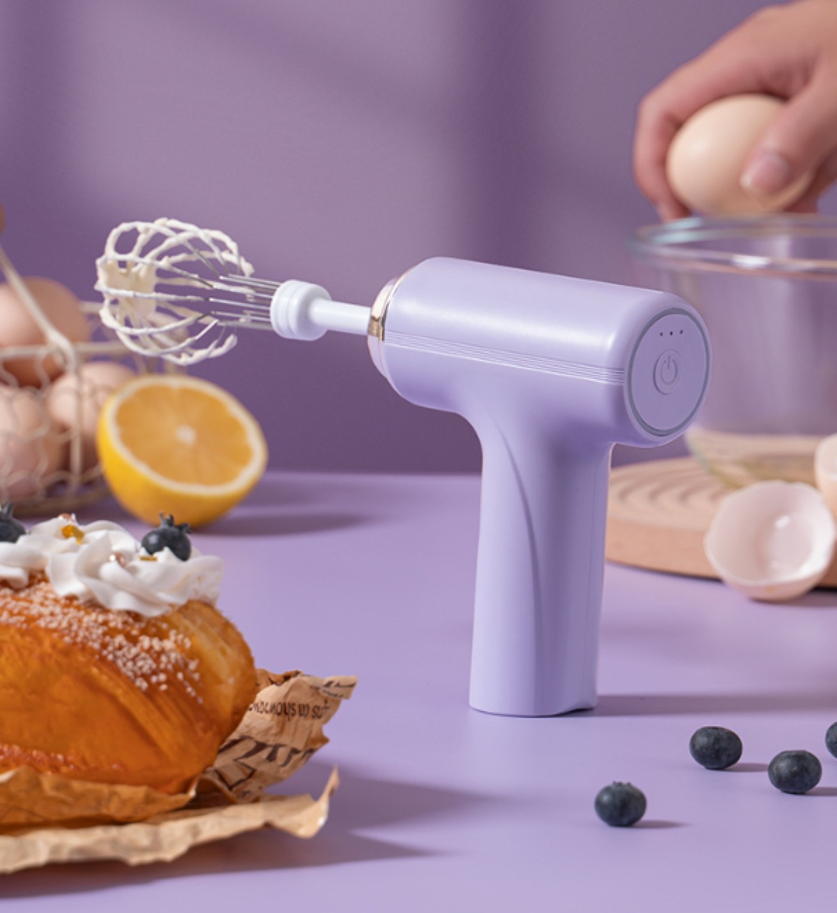 Ein lila Handmixer mit Schneebesen über Kuchen, Eiern und Blaubeeren auf einer lila Oberfläche.