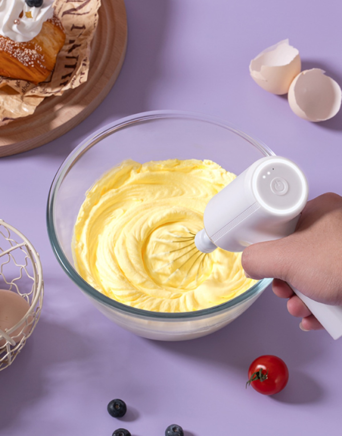 Eine Hand, die einen weißen elektrischen Mixer hält, der gelbe Creme in einer Glasschüssel verrührt. Blaubeeren und Tomaten in der Nähe.