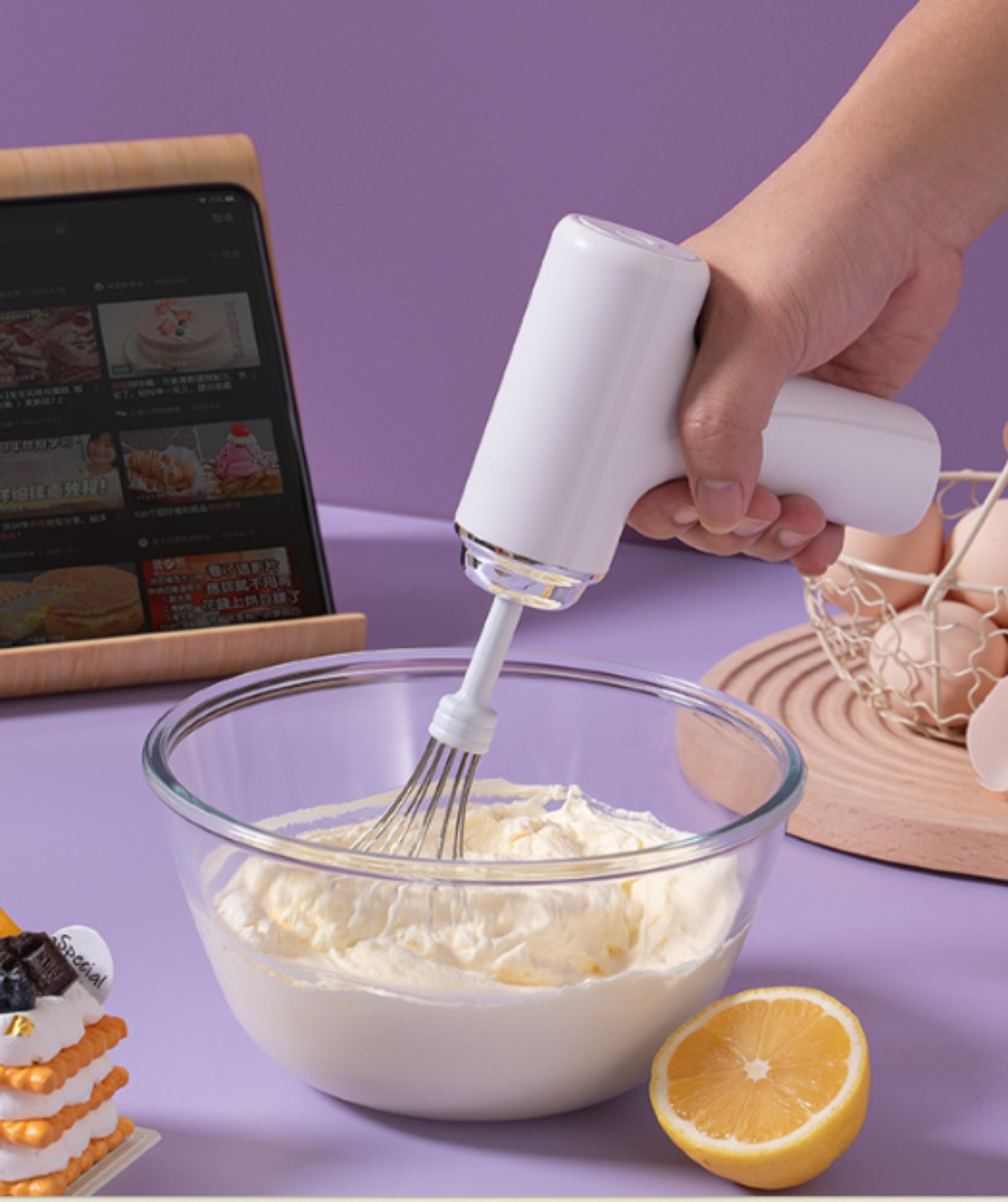 Ein Handmixer verrührt Sahne in einer Glasschüssel, ein Tablet im Hintergrund. Eier und eine Zitrone liegen daneben.