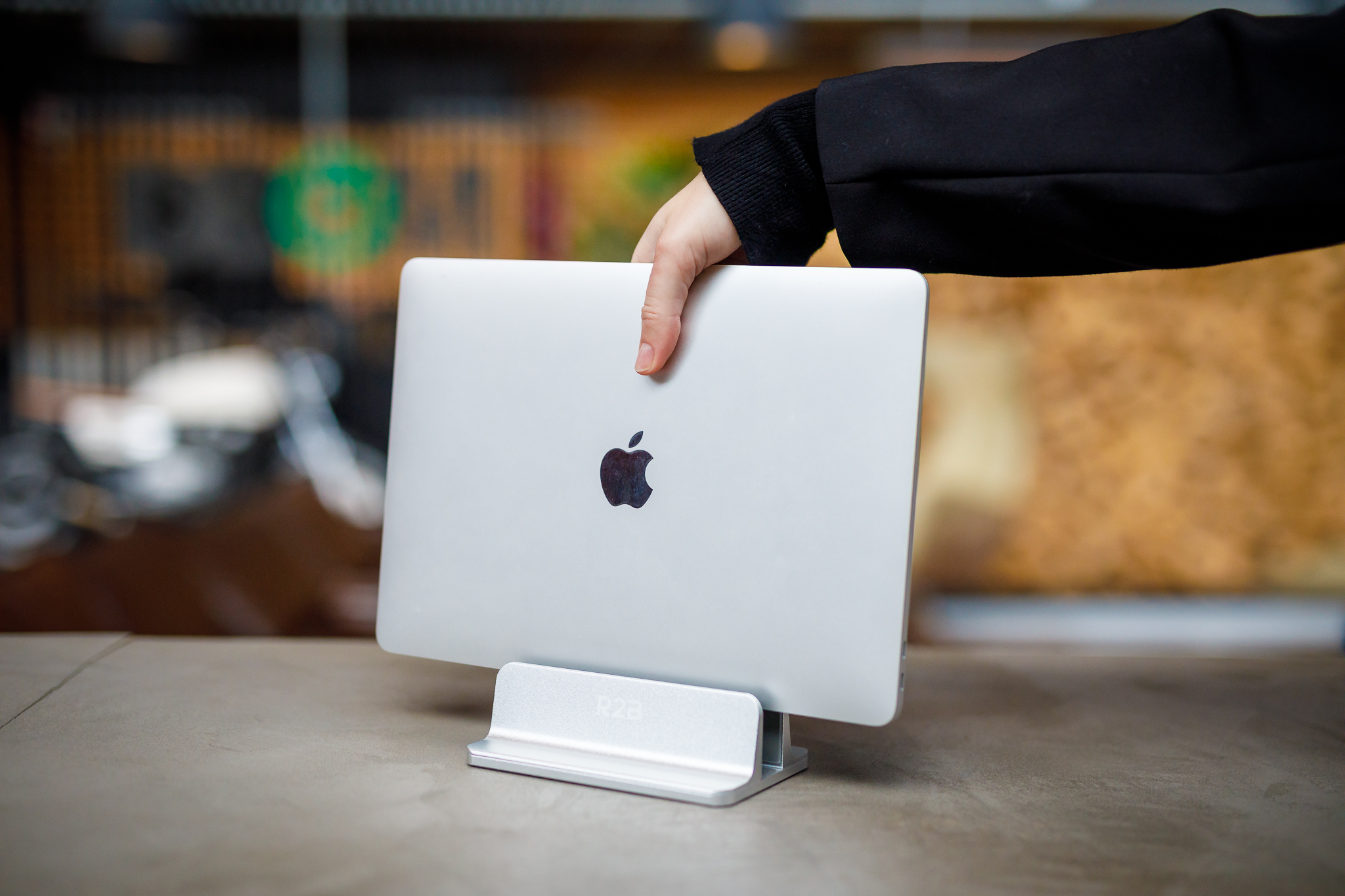 Een hand die een zilveren laptop vasthoudt in een zilveren laptopstandaard op een tafel. Apple-logo zichtbaar.