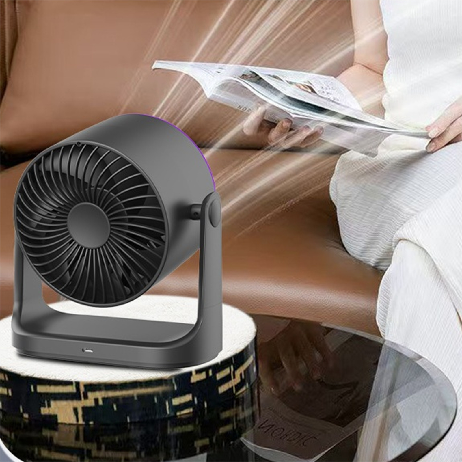 Kleiner schwarzer Ventilator auf einem Ständer, im Hintergrund liest eine Person eine Zeitschrift.