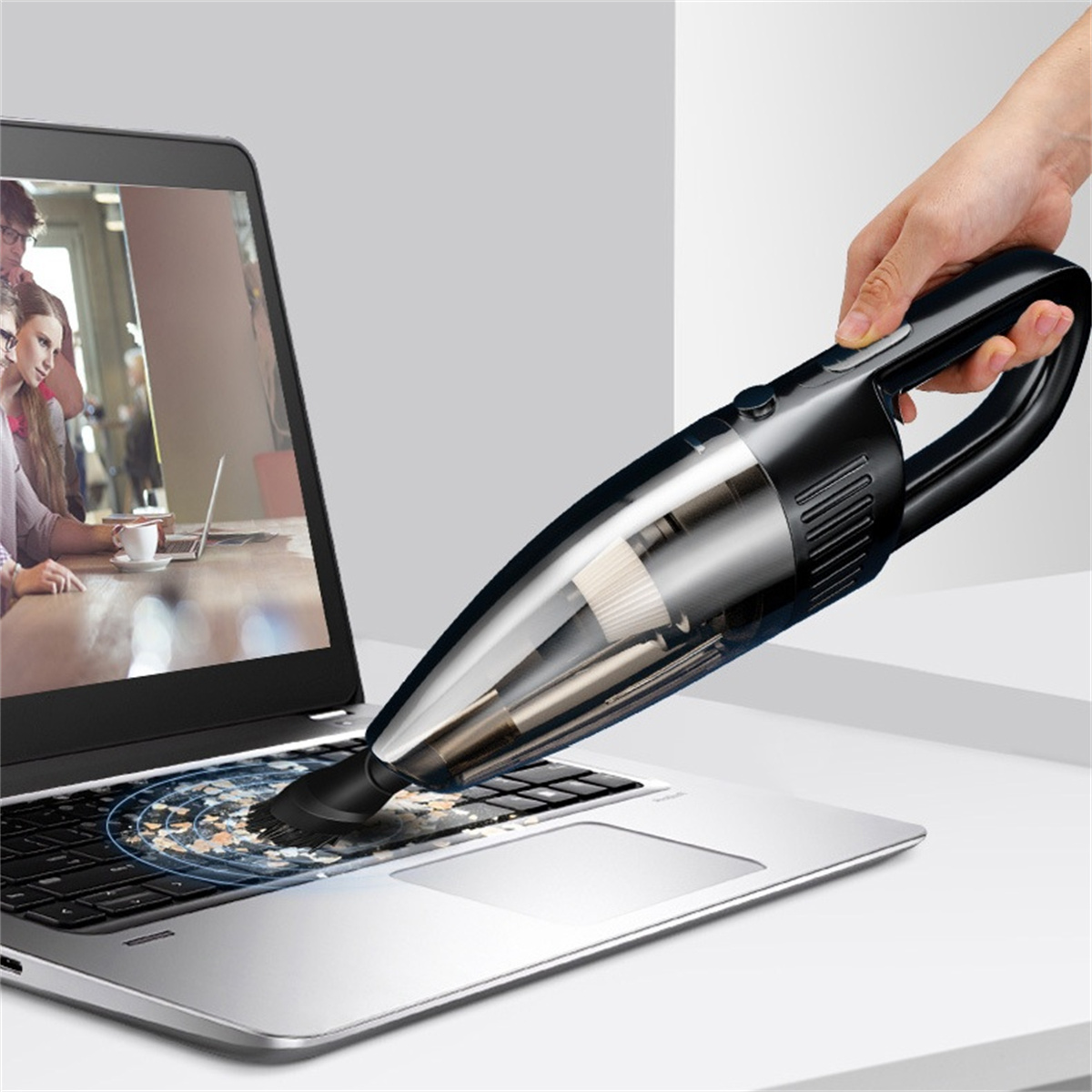 Eine Hand saugt eine Laptop-Tastatur ab und entfernt Staub. Personen im Hintergrund.