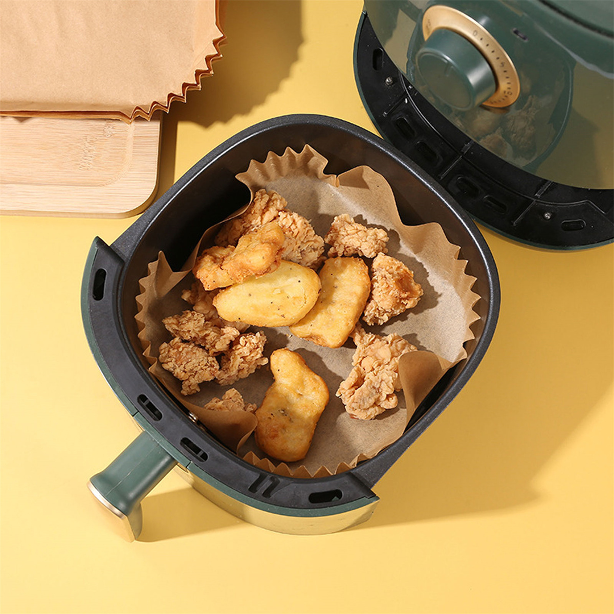 Heißluftfritteuse mit Frittiertem darin. Braunes Essen mit Papiereinlage. Grüner und gelber Hintergrund.