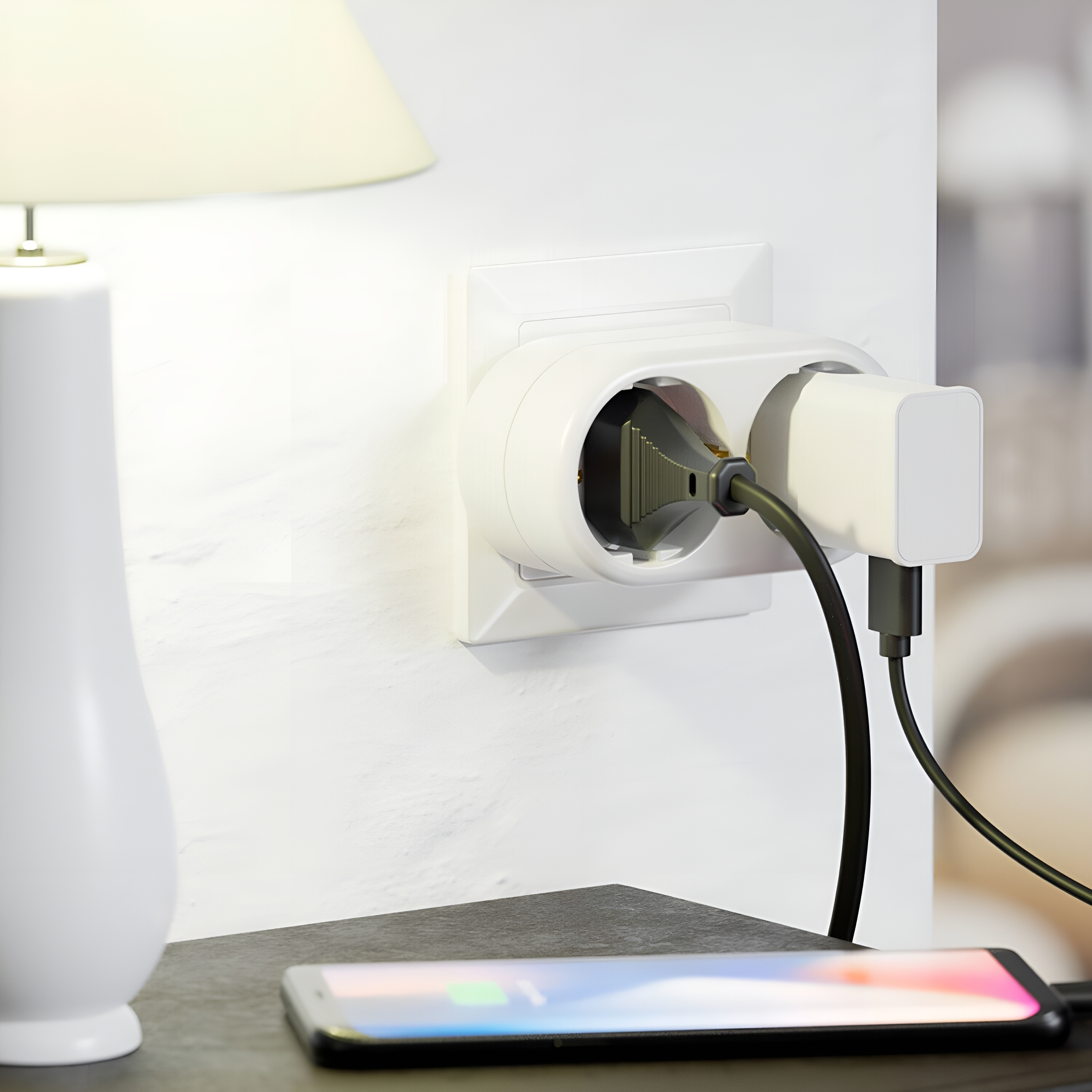 Weiße Wandsteckdose mit eingestecktem Ladegerät, ein weiteres Ladegerät und Telefon auf einem Tisch neben einer Lampe.