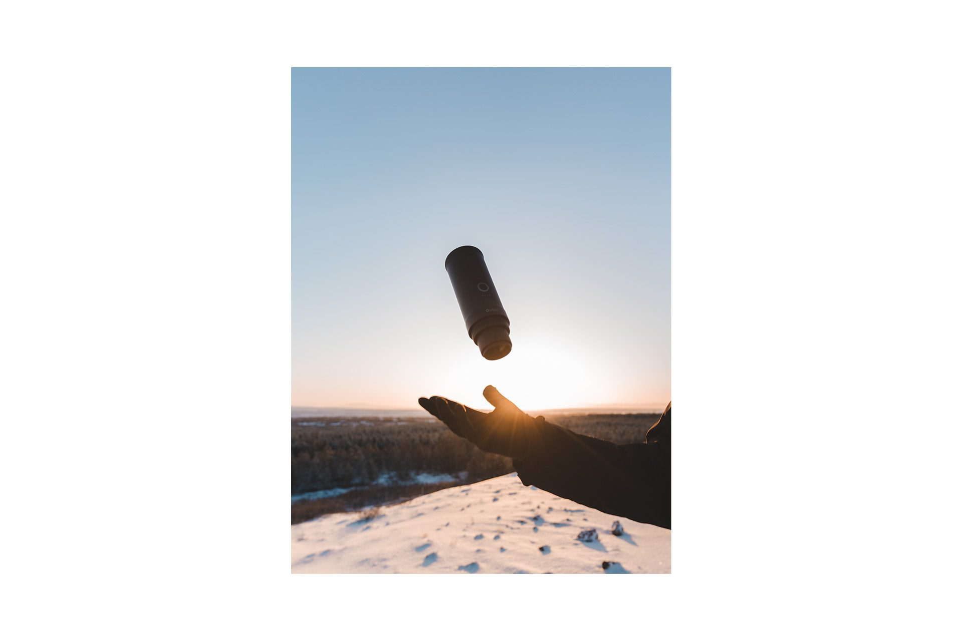 Dłoń wyrzuca ekspres do góry na tle zachodu słońca nad zaśnieżonym krajobrazem.