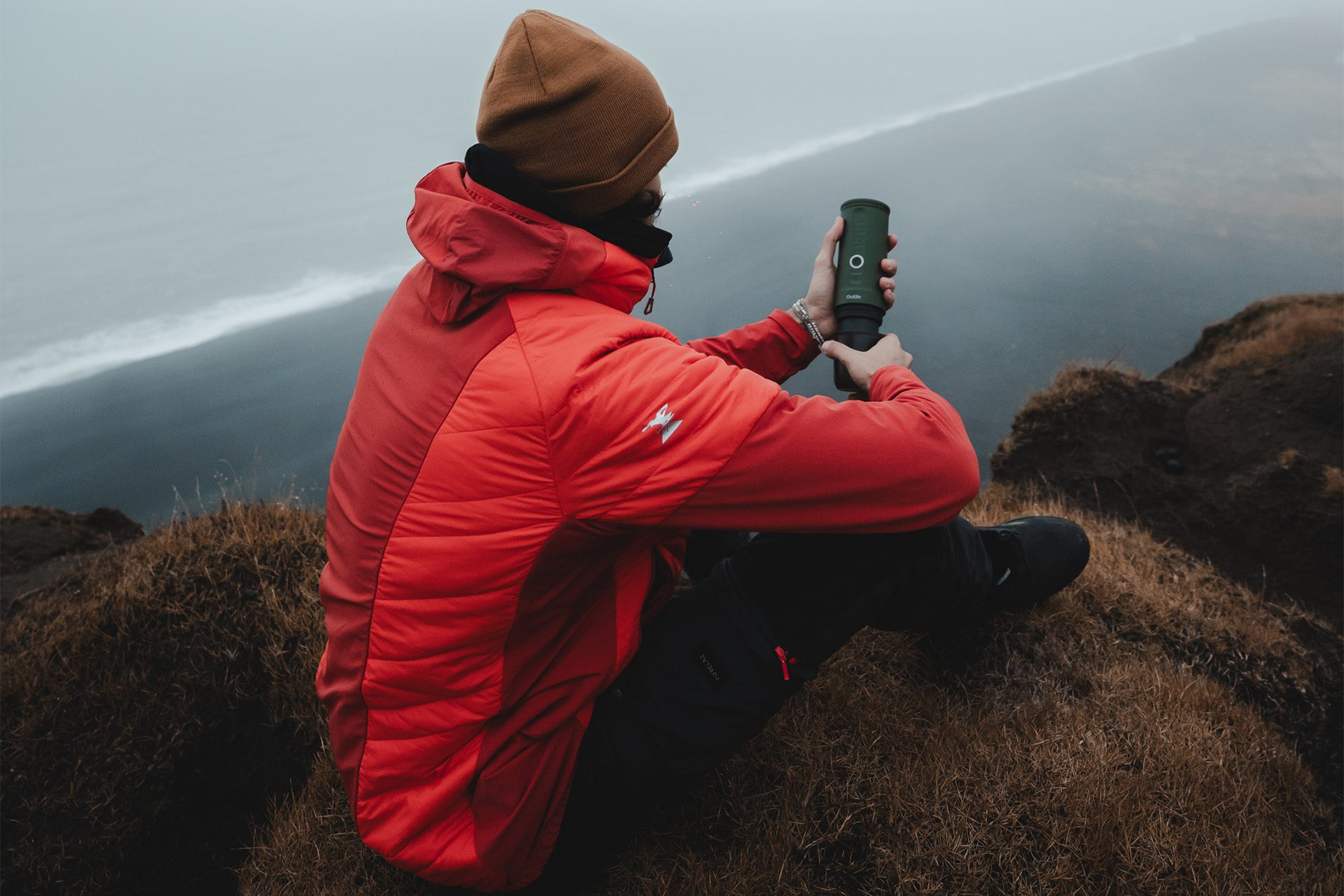 Osoba siedząca na zewnątrz trzymająca przenośny ekspres do kawy z malowniczym, mglistym tłem.