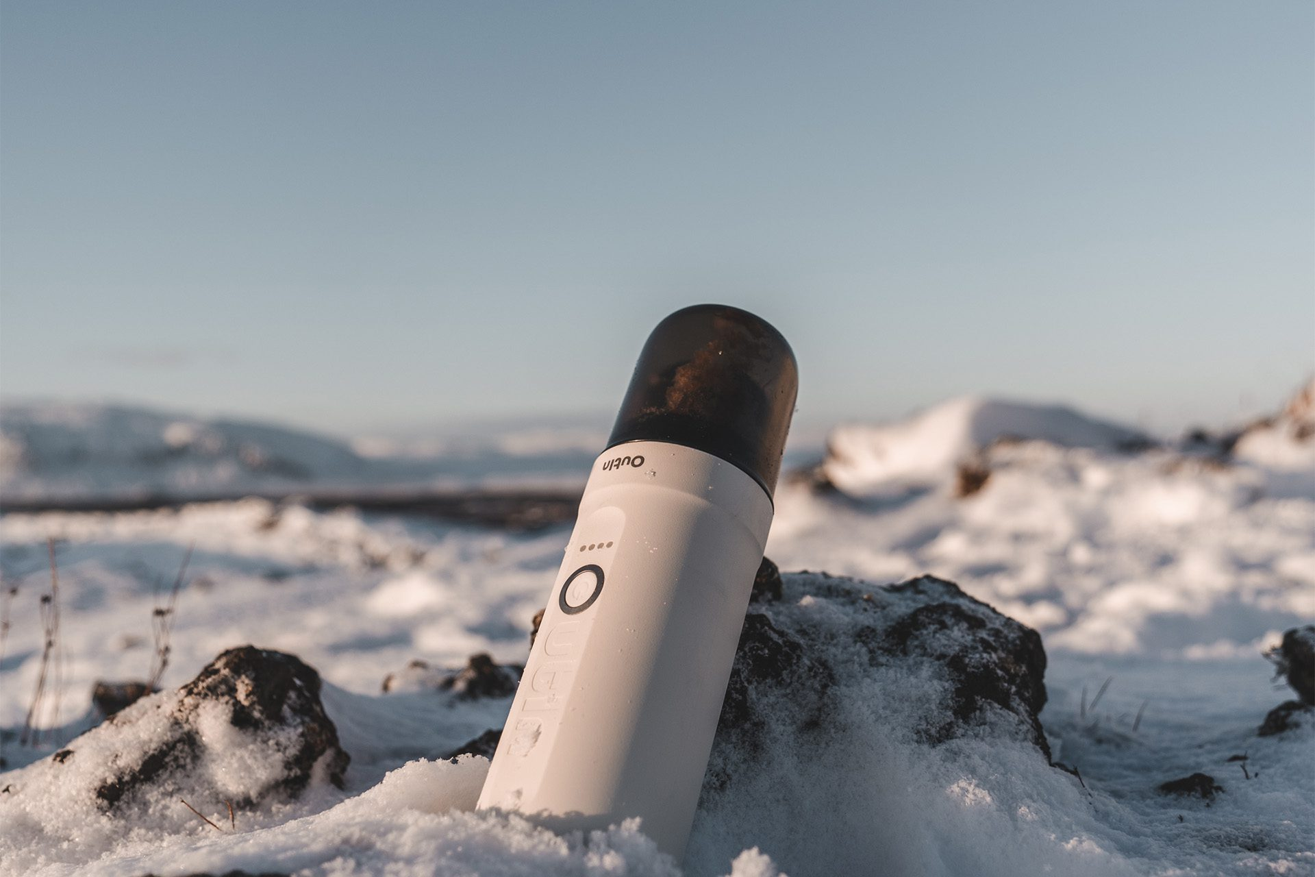Een witte koffiezetter in de sneeuw, met een donkere bovenkant, voor een besneeuwd landschap.