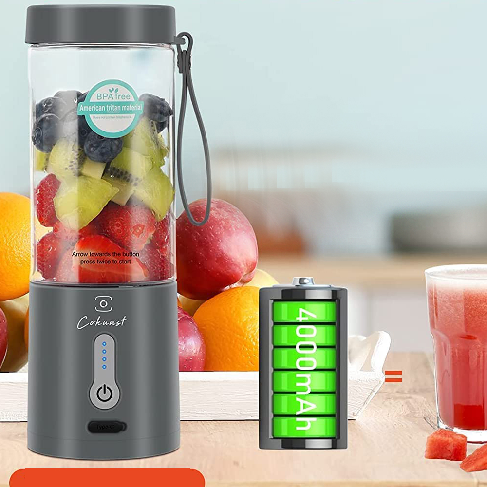 Ein grauer Mixer mit einem durchsichtigen Behälter, der mit Obst gefüllt ist. Eine Batterie und ein Glas mit Saft sind rechts.