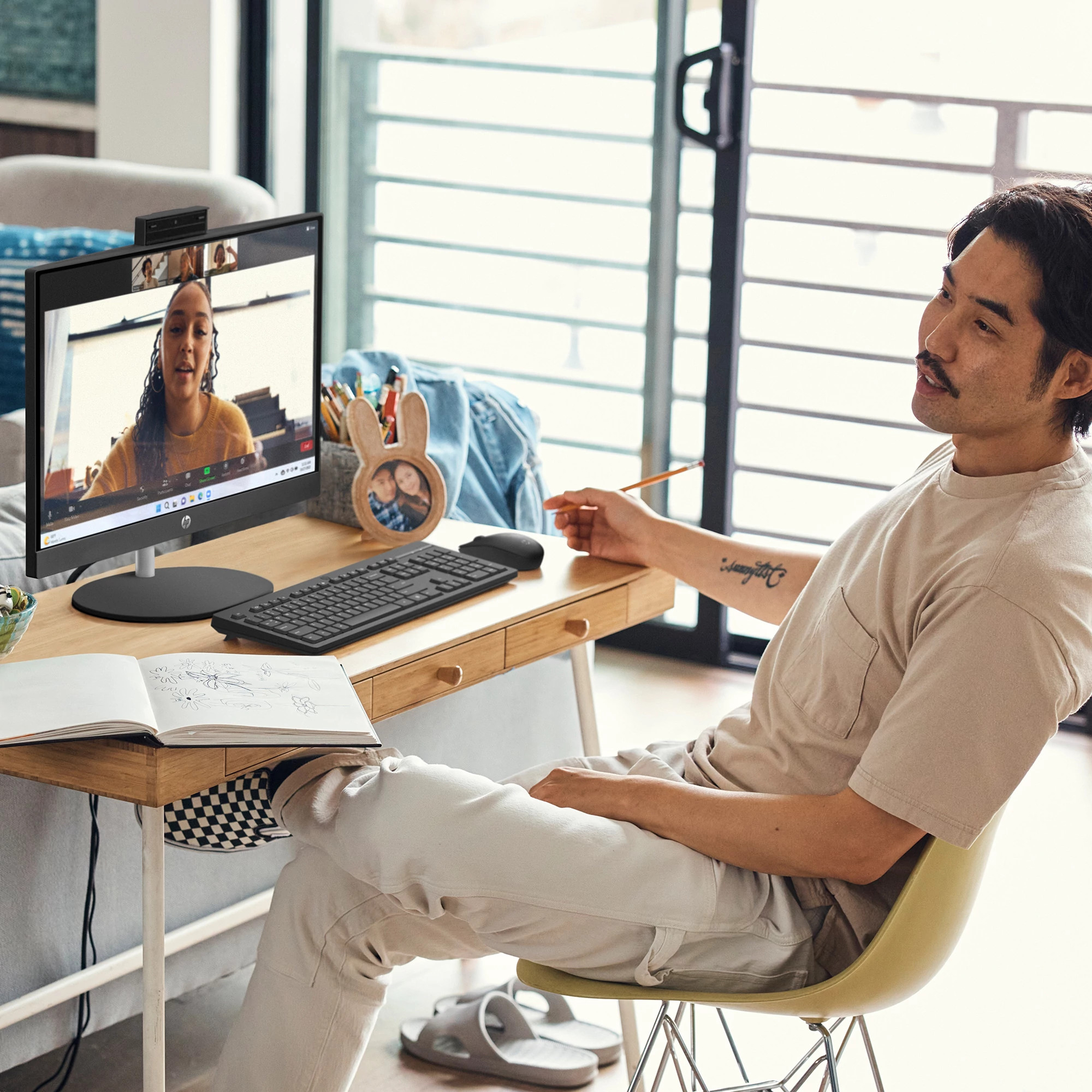 Mężczyzna na rozmowie wideo, biurko z monitorem, klawiaturą i otwartą książką. Tło z oknem.